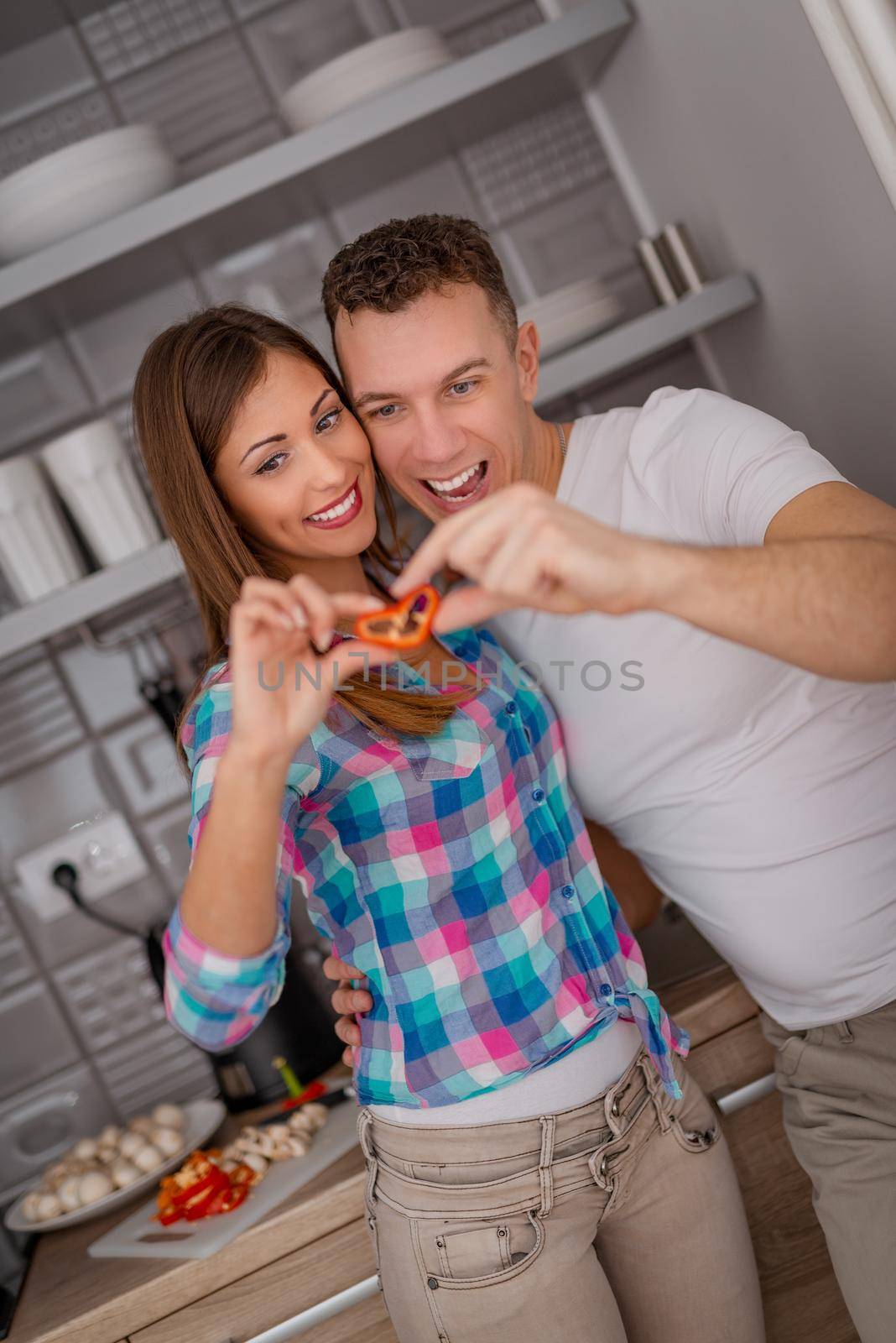 A happy young couple is having fun in the kitchen and holding a slice of red paprika in heart shape. 