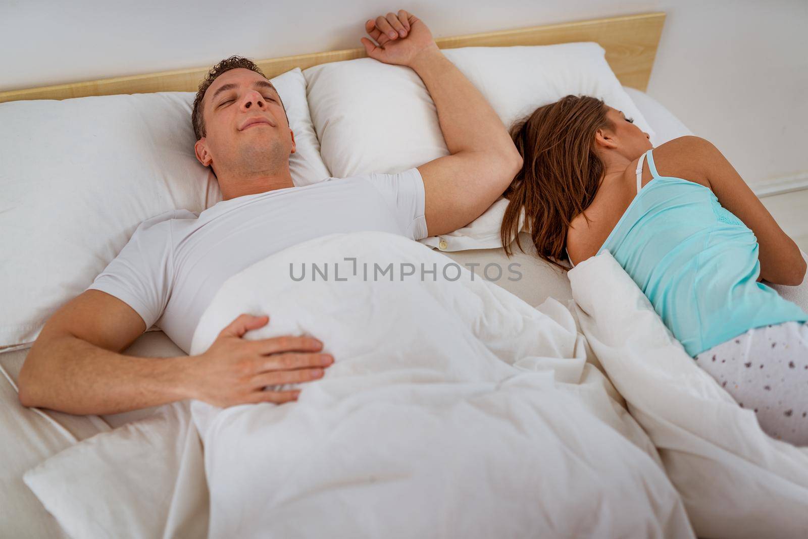 Beautiful couple sleeping together in bedroom. He sleeps relaxed and she is gathered at the end of the bed uncovered.