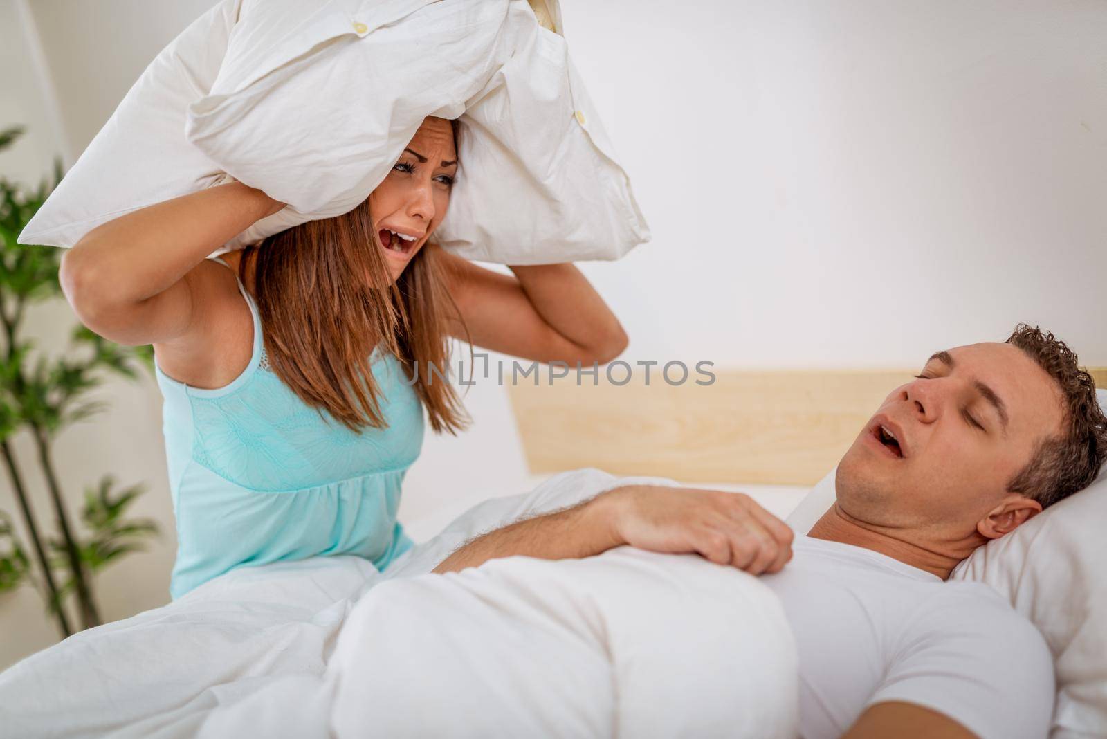 Heterosexual couple in bed, man sleeps and snoring with mouth open, while a tired woman irritated by snoring sitting on bed with a pillow on her head.