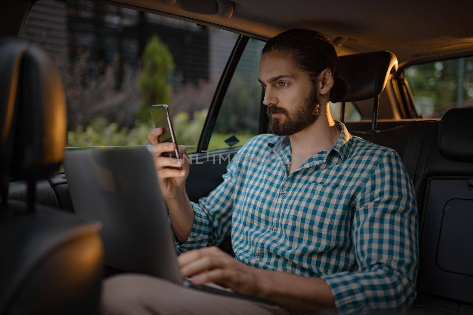 Young confident businessman is holding smartphone in his hand and texting on the back seat in car.