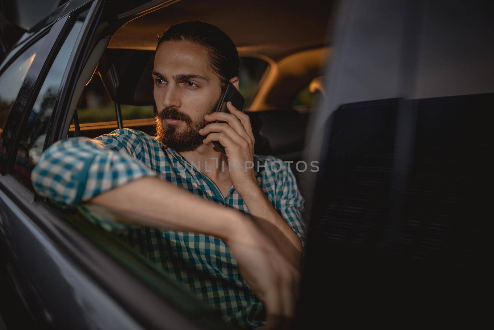 Young confident businessman is holding smartphone in his hand and talking on the back seat in car.