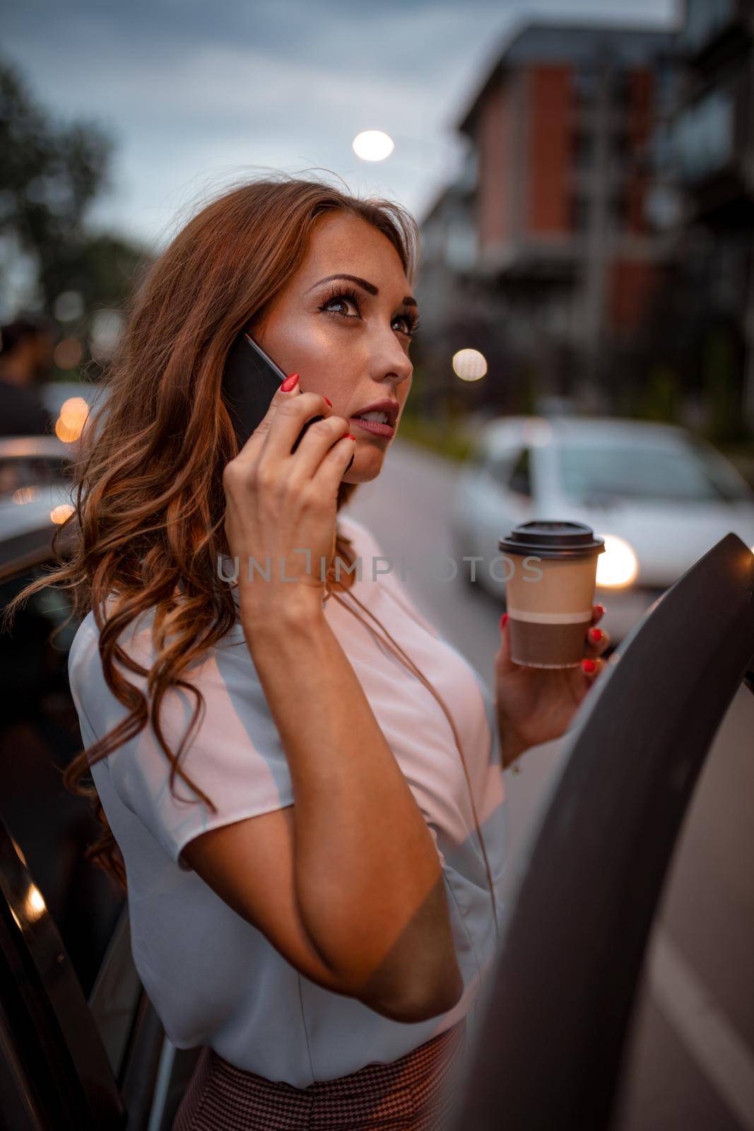 A young beautiful woman is talking on the smartphone holding a cup of coffee, and is going to enter in the car.
