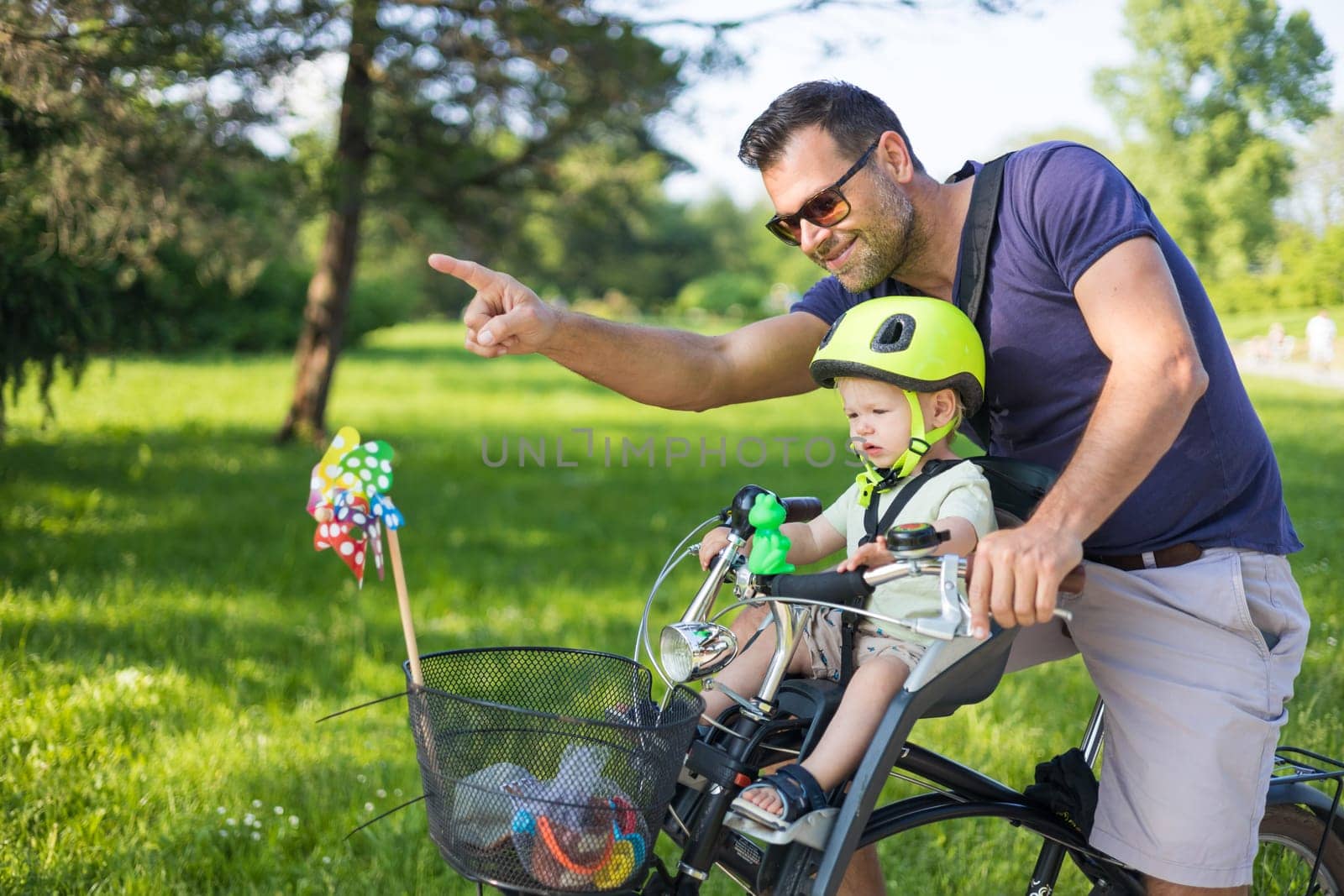 Look over there. Active family day in nature. Father and son ride bike through city park on sunny summer day. A cute boy is sitting in front bicycle chair while father rides bicycle. Father son bonding. by kasto