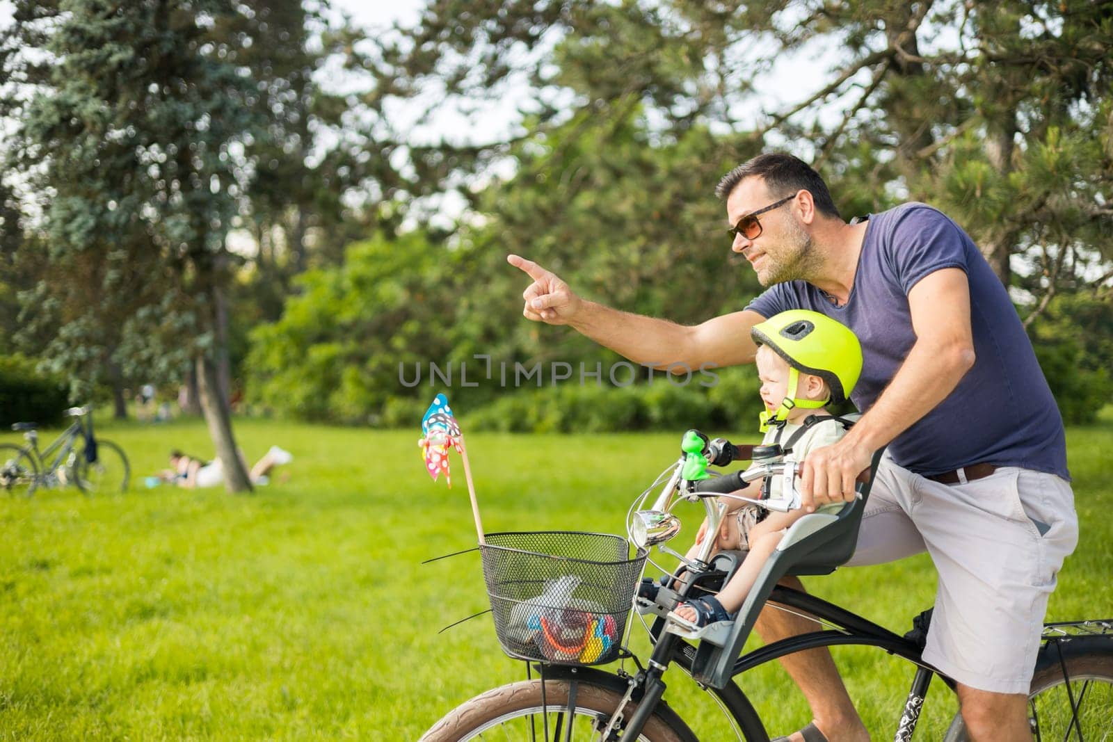 Look over there. Active family day in nature. Father and son ride bike through city park on sunny summer day. A cute boy is sitting in front bicycle chair while father rides bicycle. Father son bonding. by kasto