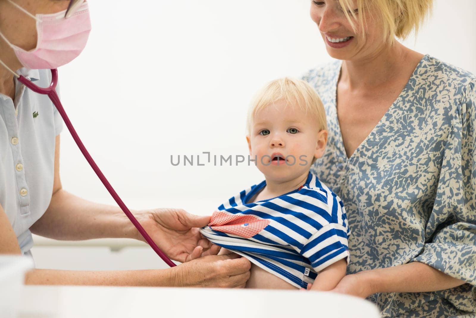 Infant baby boy child being examined by his pediatrician doctor during a standard medical checkup in presence and comfort of his mother. National public health and childs care care koncept