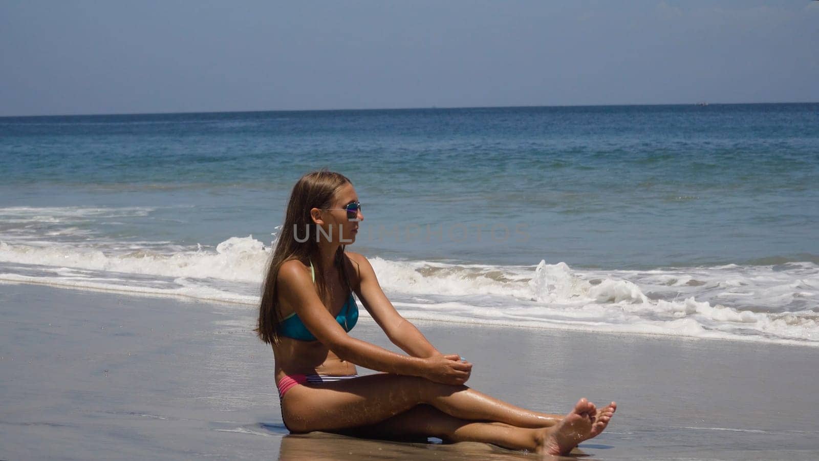 Girl in the sun glasses sits on a sandy beach among the waves and looks at the sea.Young girl relaxing at Tropical Beach. Recreation and pampering on the seashore ocean, river, lake in summer and sunny day.