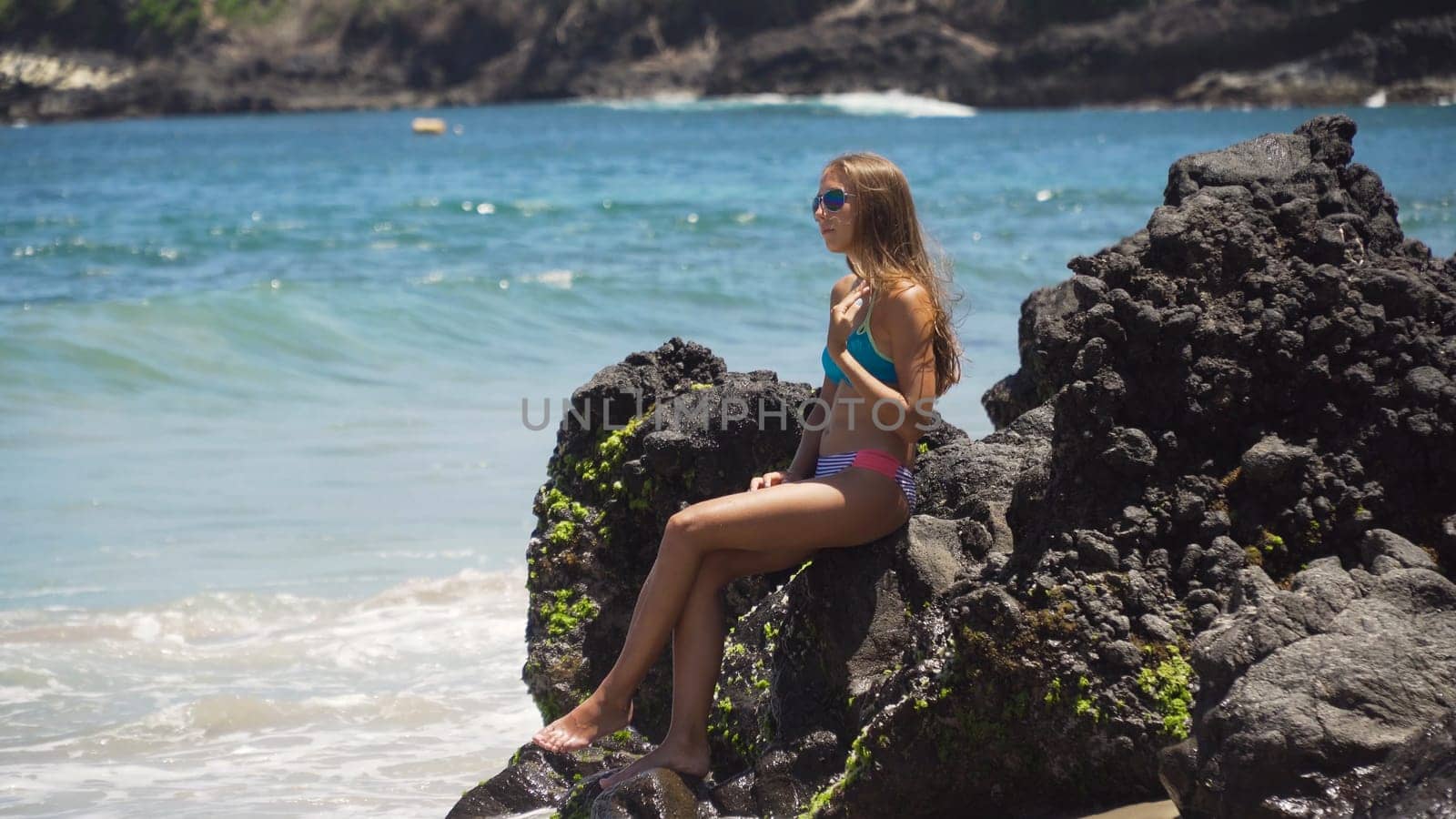 Beautiful girl in bikini with fluttering hair sitting on a rock above the sea and looking at ocean. Girl on the tropical beach, Bali, Indonesia. Travel concept, Travel concept.