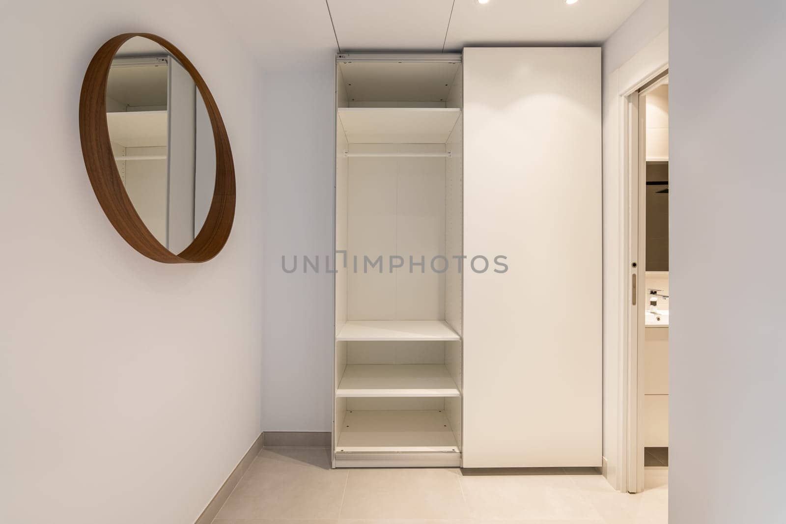 Narrow room with mirror and an open wardrobe with an entrance to the bathroom in light beige tones. Concept of comfortable compact apartment in a new building. Copyspace.