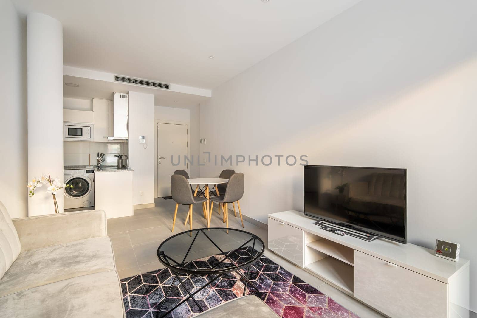 Studio apartment with a combined kitchen and living room with a corner sofa, tables and chairs and a TV with a picture on the background of a kitchen set and a hallway.
