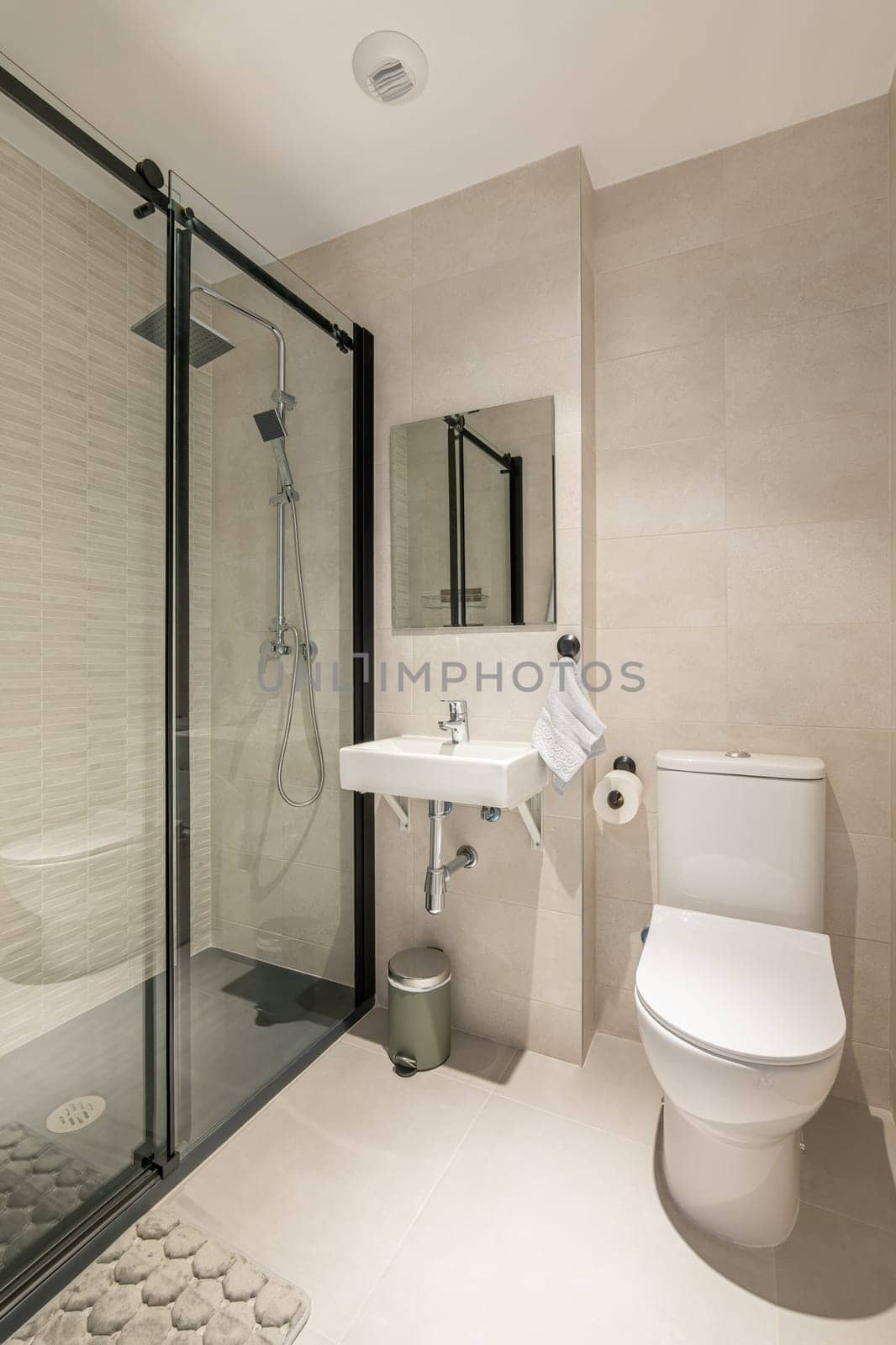 Vertical shot of comfortable bathroom with a glazed shower, toilet and sink and a mirror in light colors with beige tiles. Concept of a bathroom in a hotel with a modern interior.