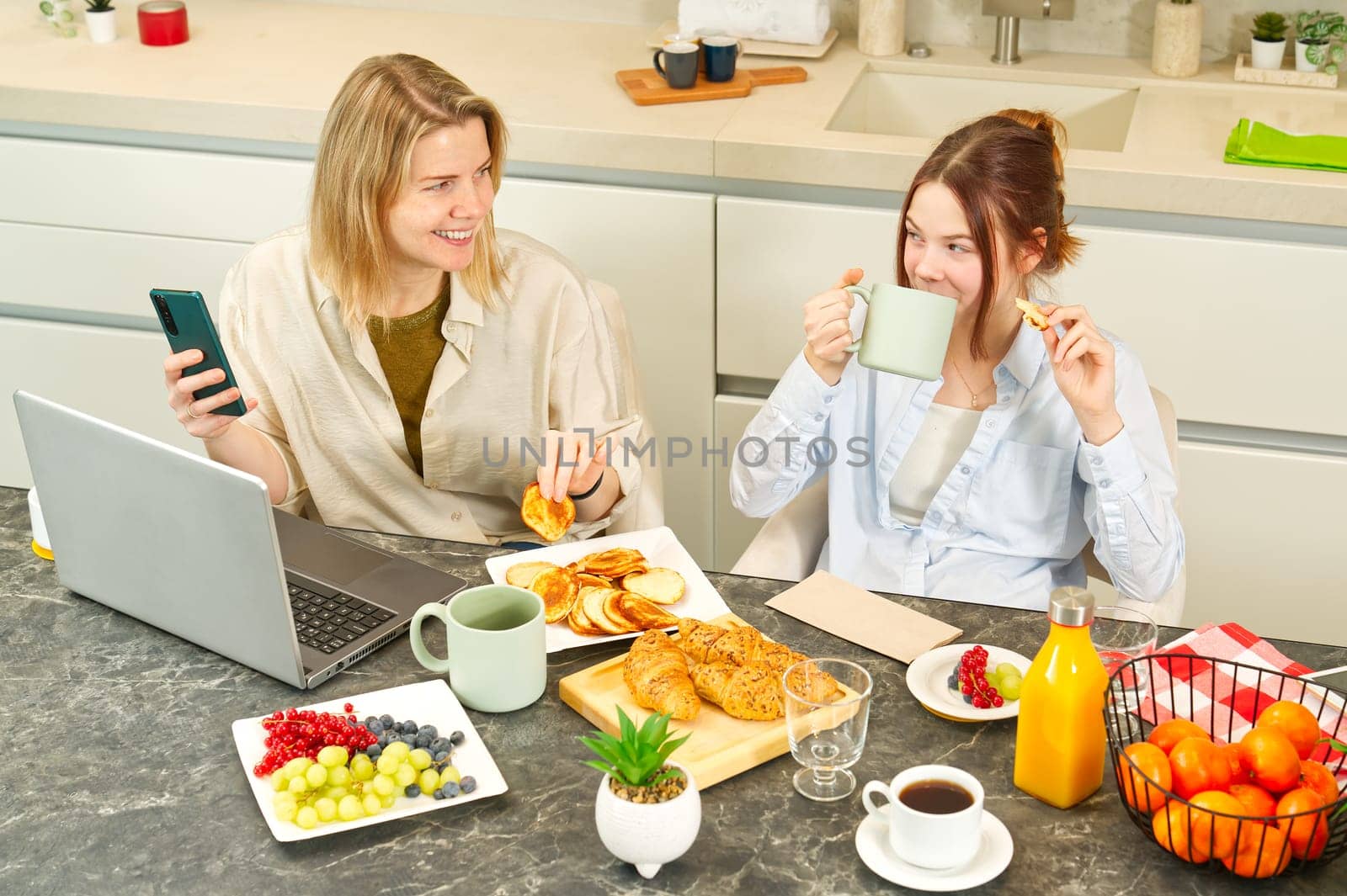 Happy mother and daughter having breakfast in kitchen and using digital devices. Lifestyle by PhotoTime