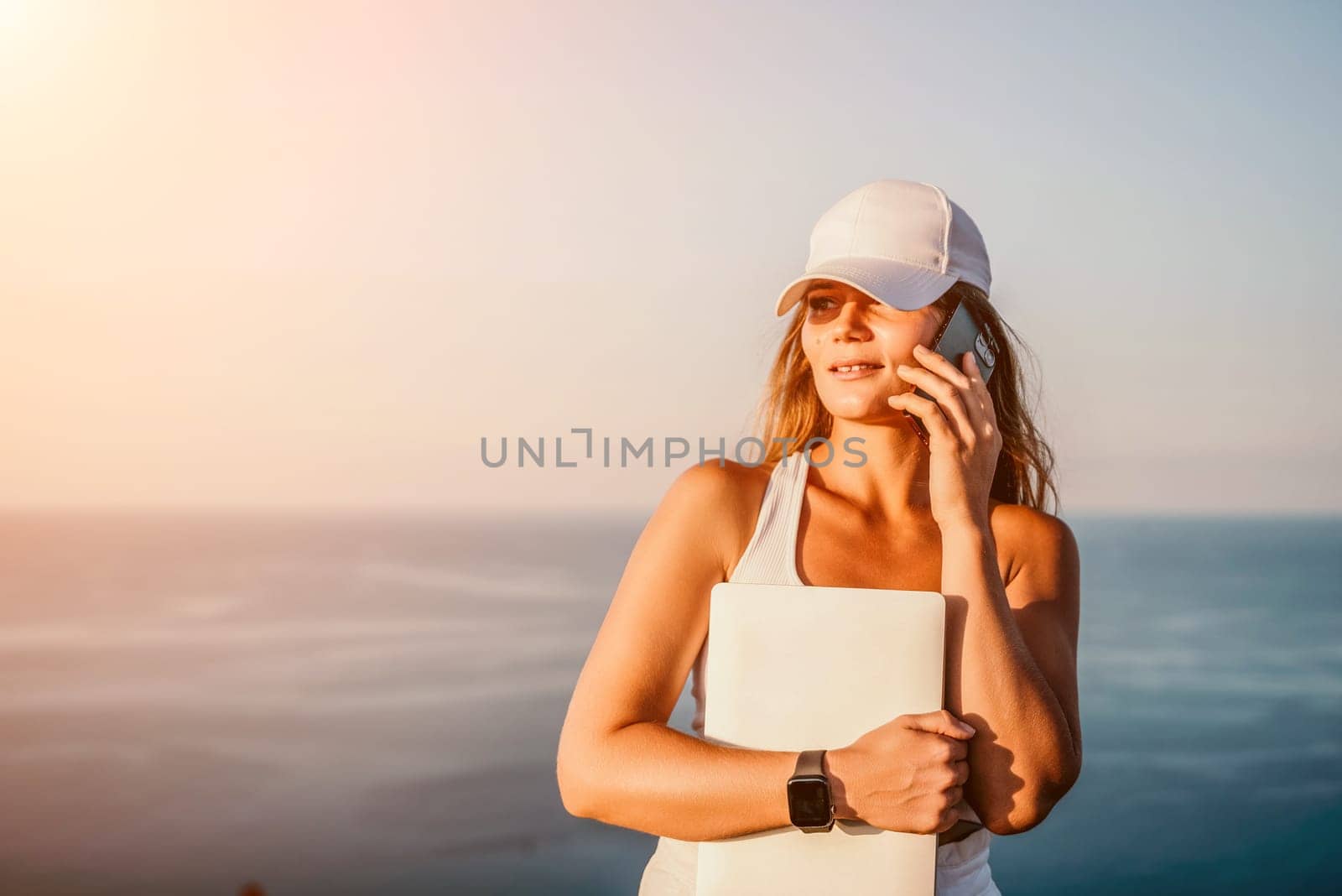 Successful business woman in yellow hat working on laptop by the sea. Pretty lady typing on computer at summer day outdoors. Freelance, travel and holidays concept.