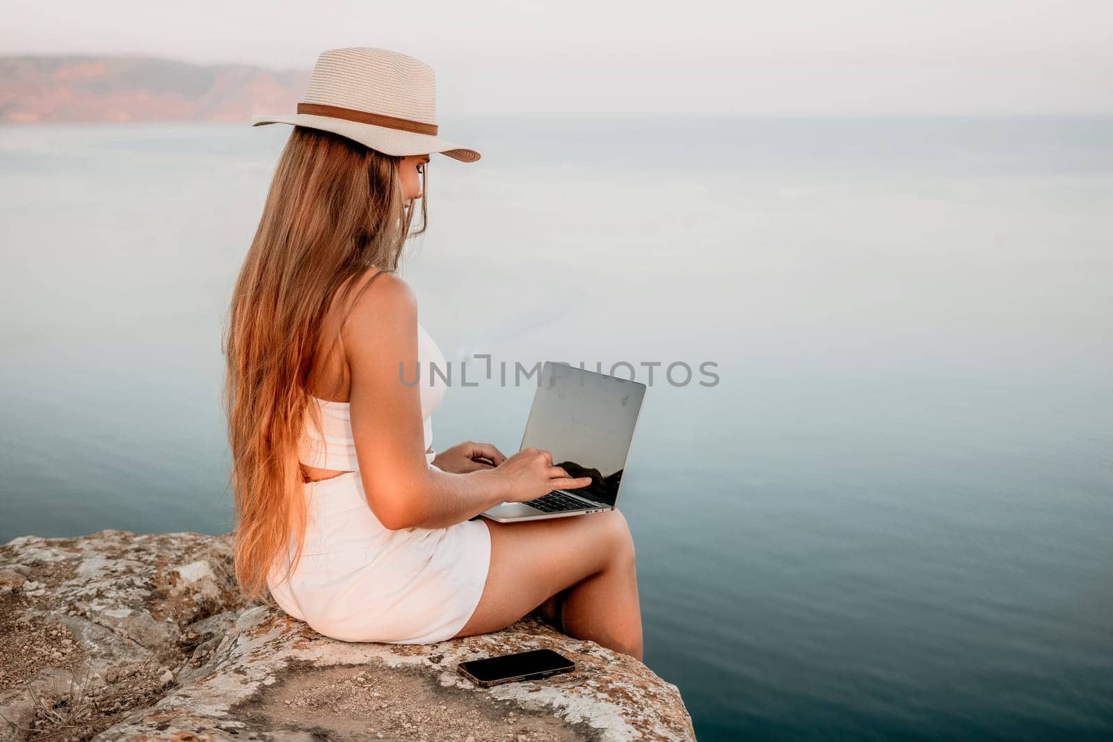 Digital nomad, Business woman working on laptop by the sea. Pretty lady typing on computer by the sea at sunset, makes a business transaction online from a distance. Freelance, remote work on vacation