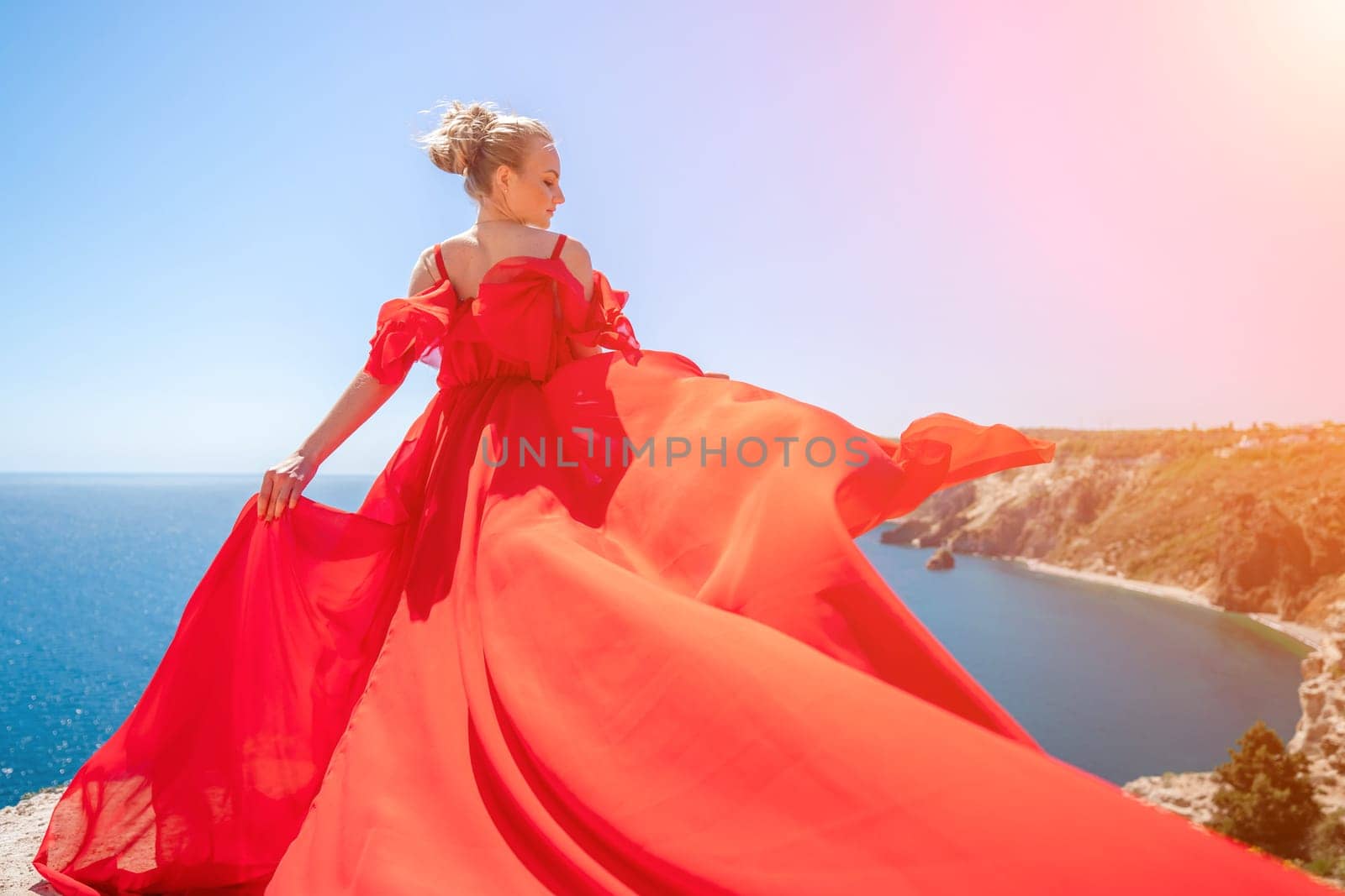 woman sea red dress. Blonde with long hair on a sunny seashore in a red flowing dress, back view, silk fabric waving in the wind. Against the backdrop of the blue sky and mountains on the seashore
