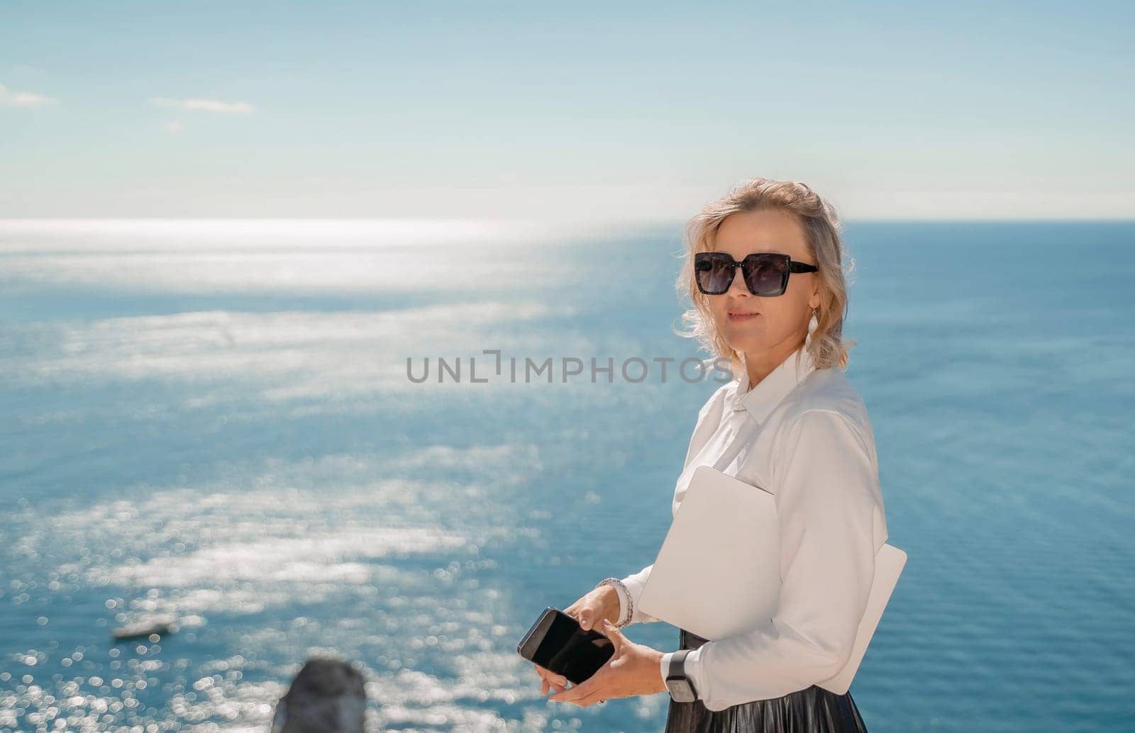 Freelance women sea working on a computer. Pretty middle aged woman with computer and phone outdoors with beautiful sea view. The concept of remote work