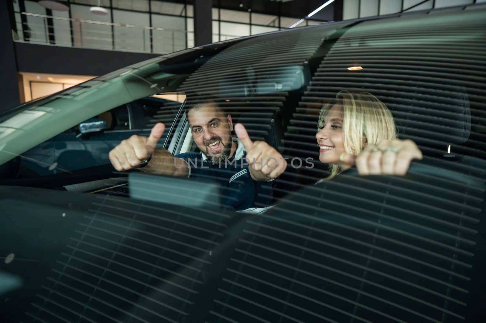 Happy caucasian couple is sitting in a new car in a car dealership. Man showing thumbs up. by mrwed54