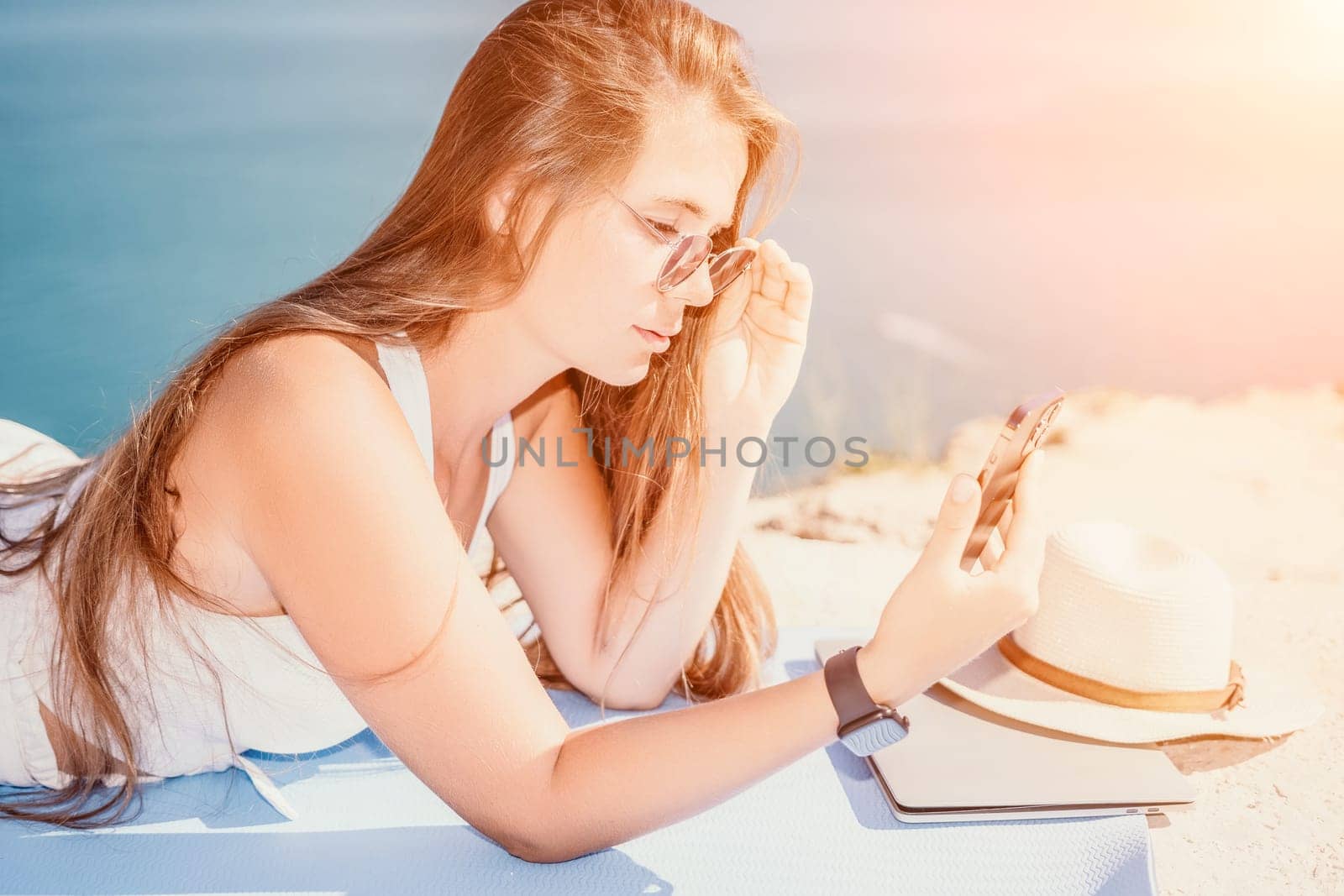 Digital nomad, woman in the hat, a business woman with a laptop sits on the rocks by the sea during sunset, makes a business transaction online from a distance. Freelance, remote work on vacation. by panophotograph