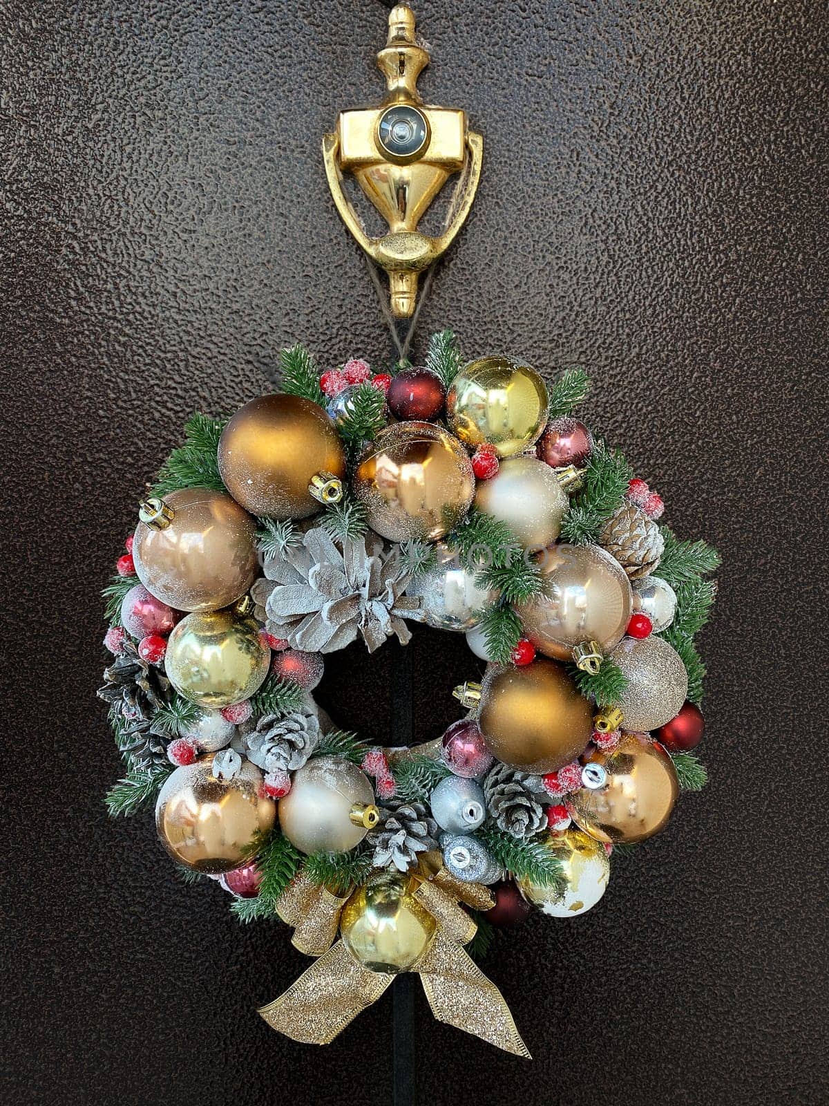 Luxurious Christmas decorative wreath, handmade with pine cones and golden balls, hangs on the dark front door of the house.