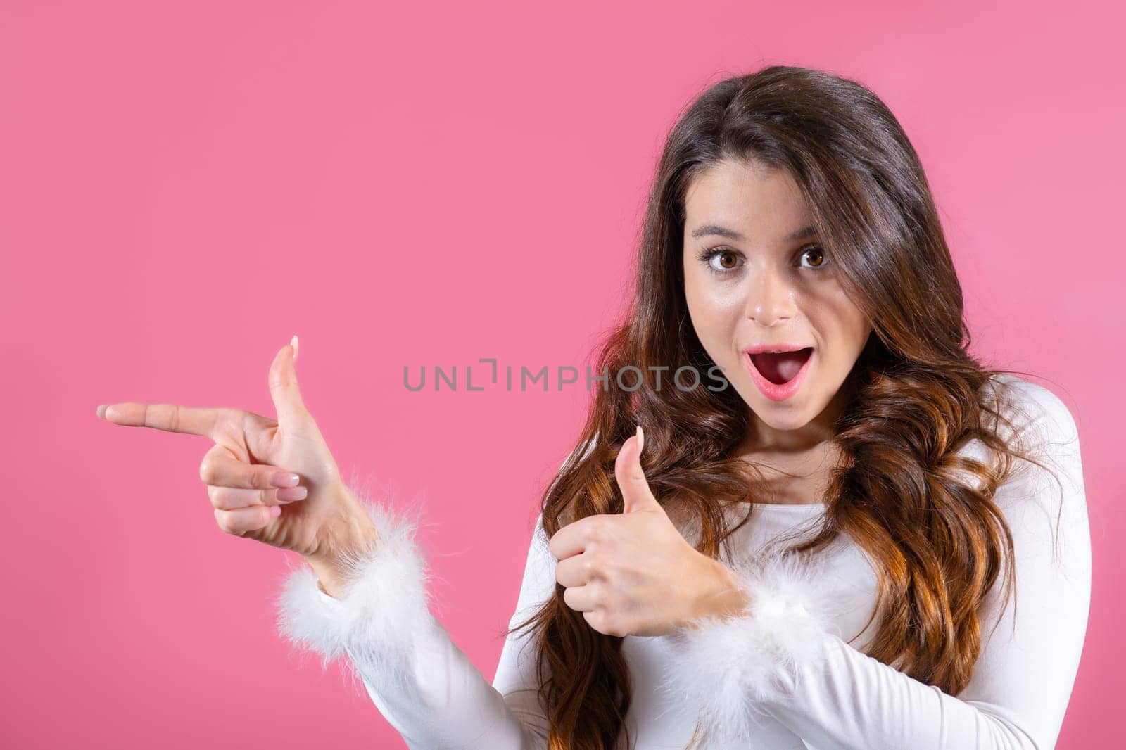 A portrait of a young lady looks at the camera and stands on a pink backdrop, gesturing towards copy space.