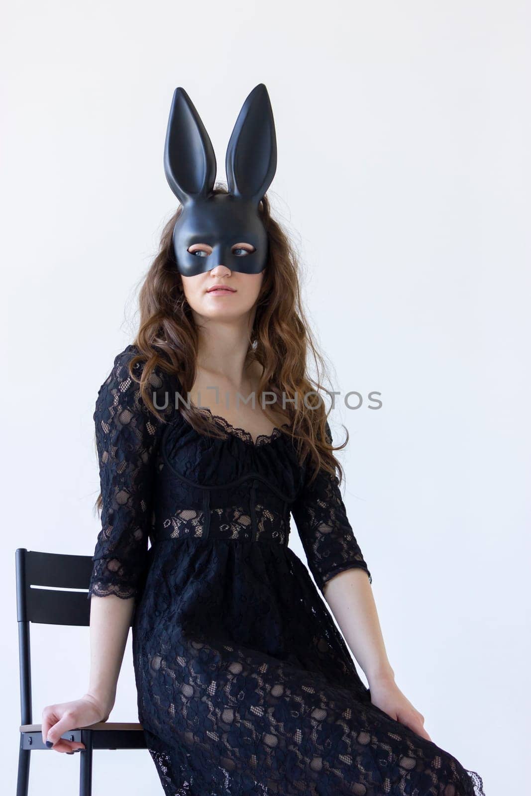 Young Woman Wearing Rabbit, Hare Black Mask On Face Sitting on chair white background, wall. Vertical Plane. High quality photo