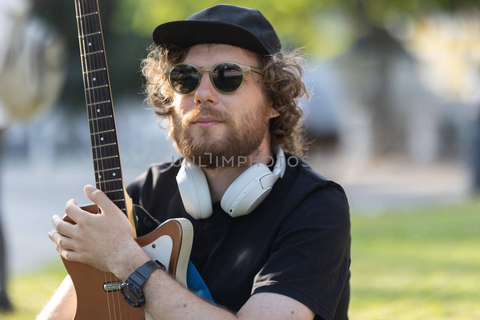 A man hipster wearing sunglasses and wireless headphones sitting in the park with his guitar . Portrait