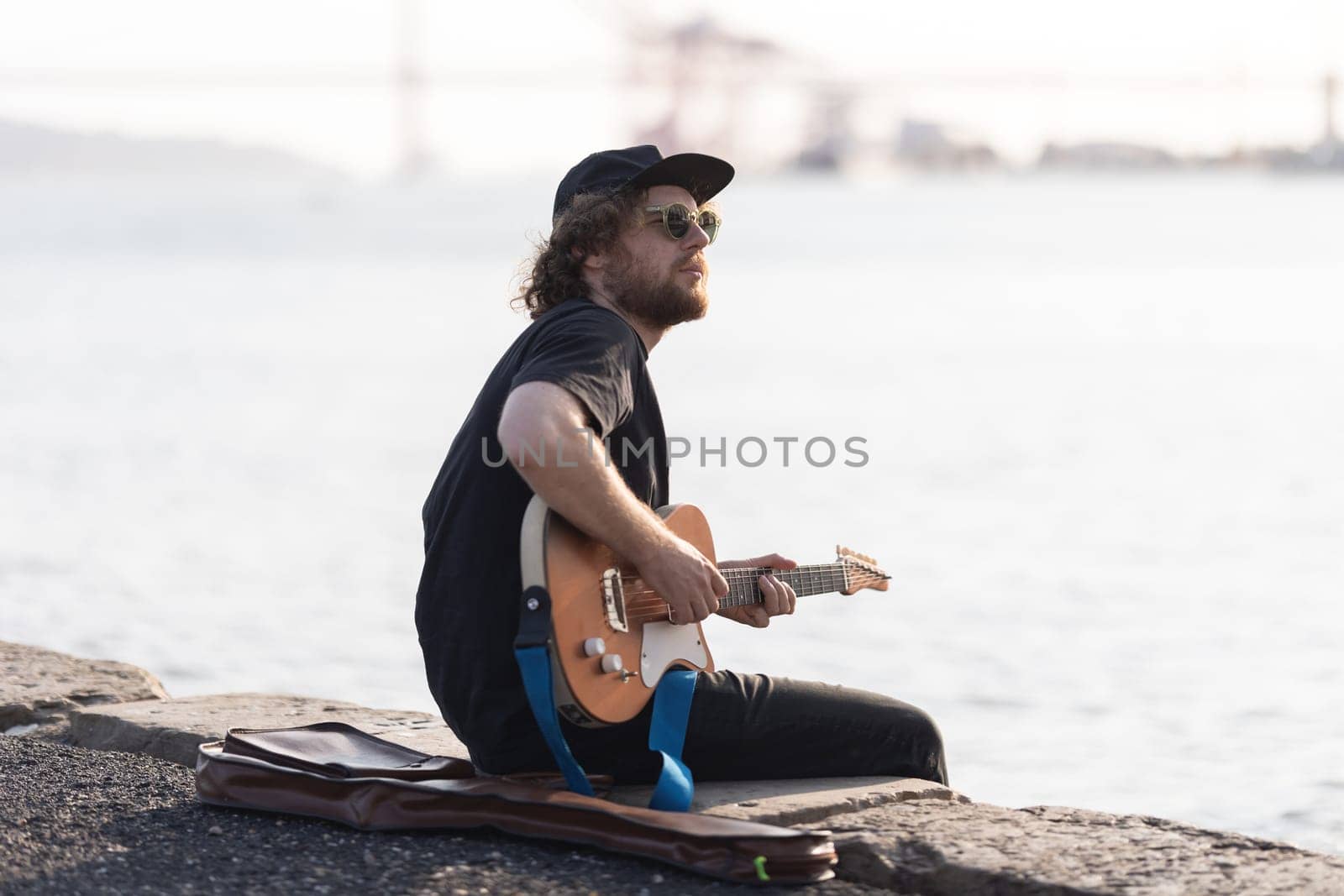 A man hipster playing electric guitar at the waterfront. Mid shot