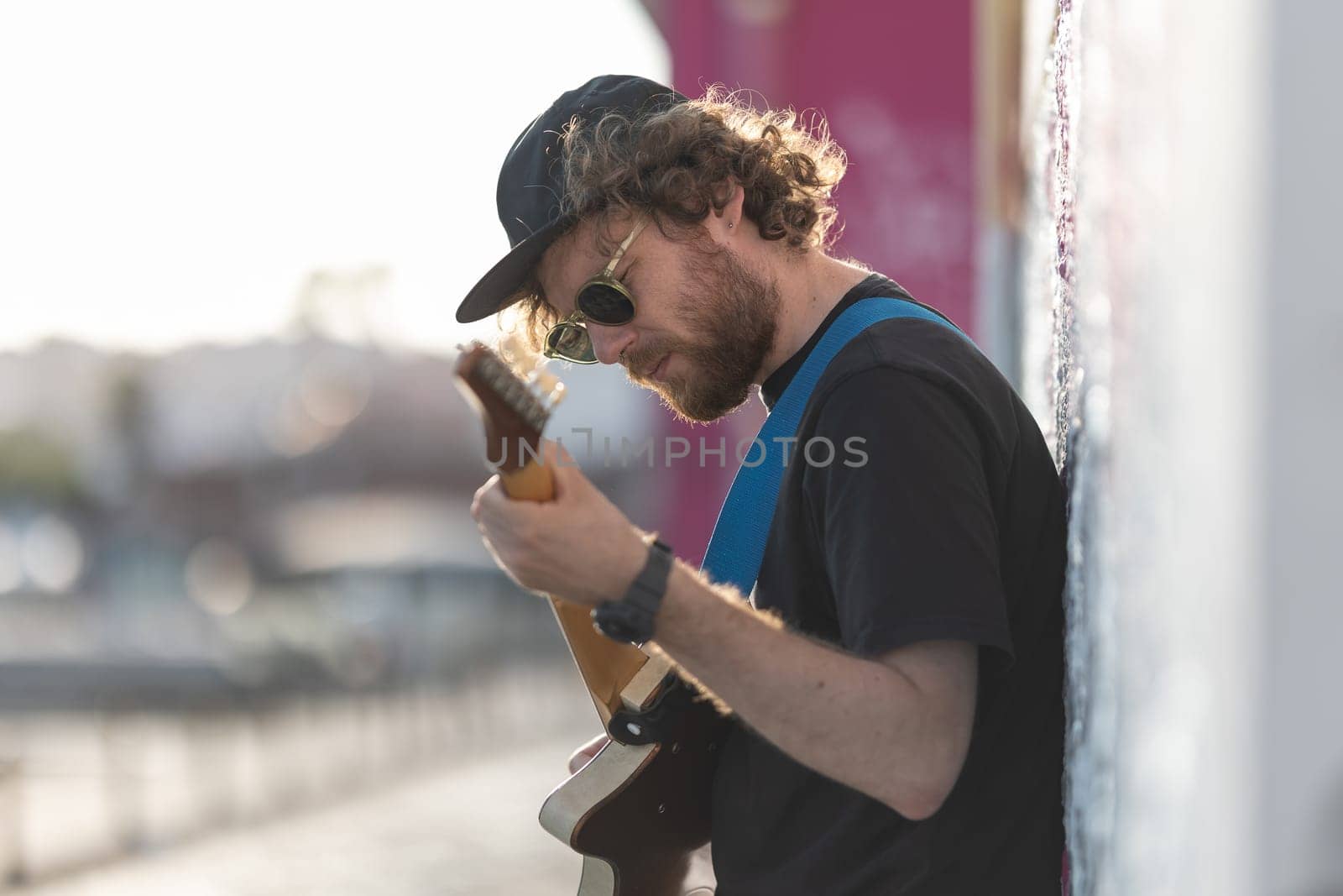 A man hipster standing by the wall and playing guitar. Mid shot