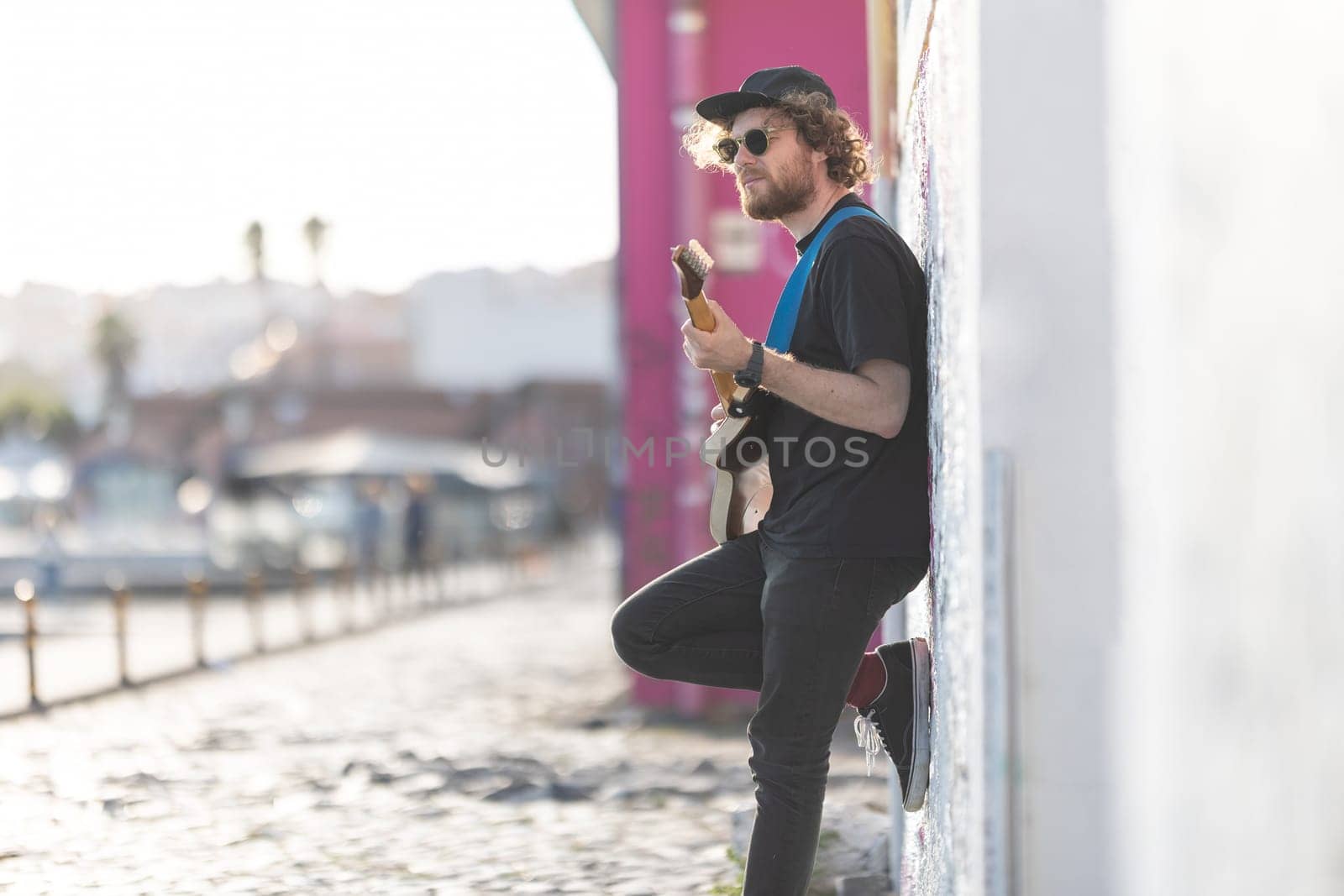 A man hipster standing by the wall and playing electric guitar. Mid shot