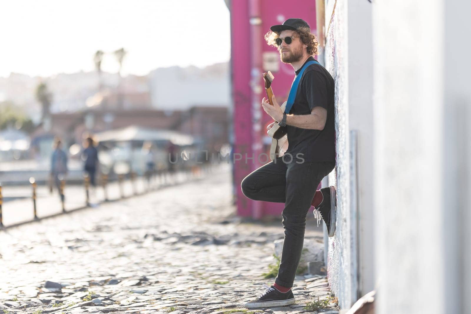 A man hipster standing by the wall and playing electric guitar on the street. Mid shot