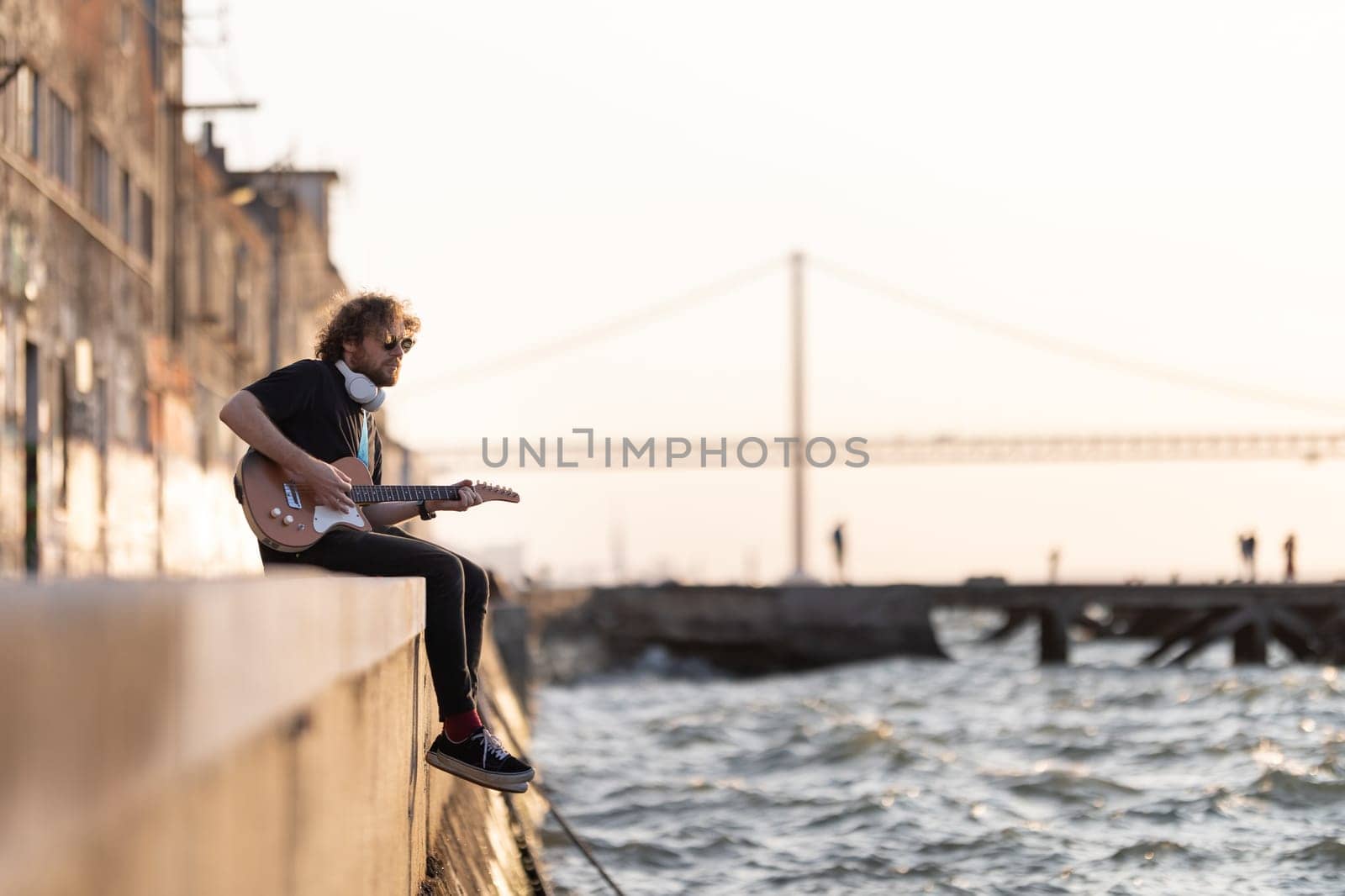 A man hipster sits on the embankment with his legs dangling to the water and plays the guitar. Mid shot