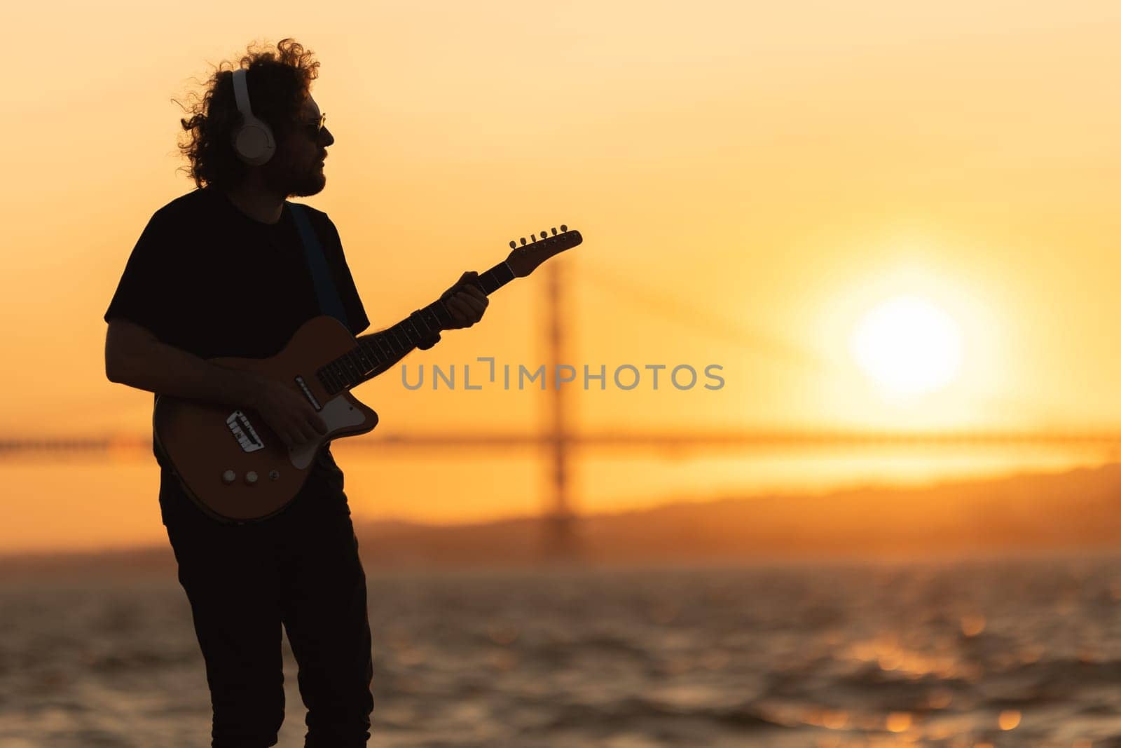 Romantic man wearing wireless headphones stands at the waterfront and playing guitar at bright orange sunset by Studia72
