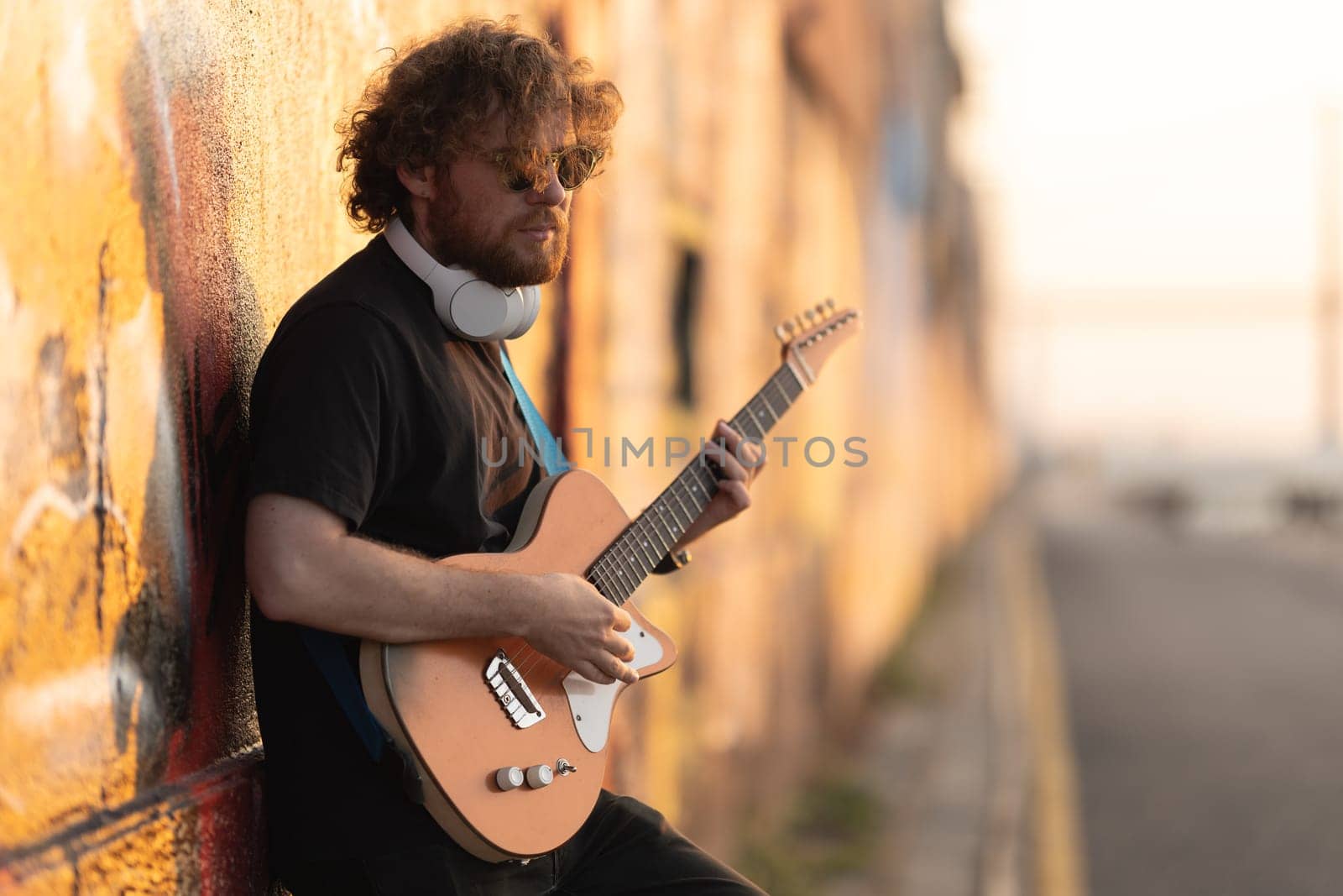 Romantic man hipster wearing sunglasses standing by the wall and playing guitar at early sunset by Studia72