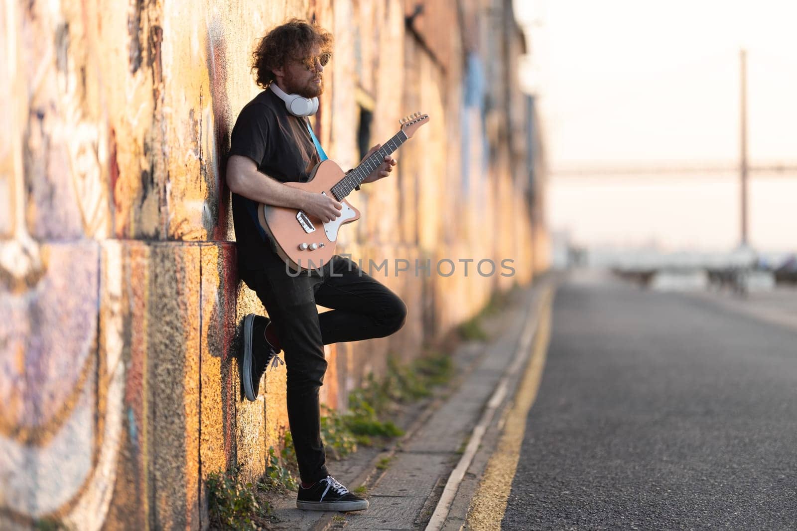 Romantic man hipster wearing sunglasses standing by the wall and playing electric guitar at early sunset. Mid shot