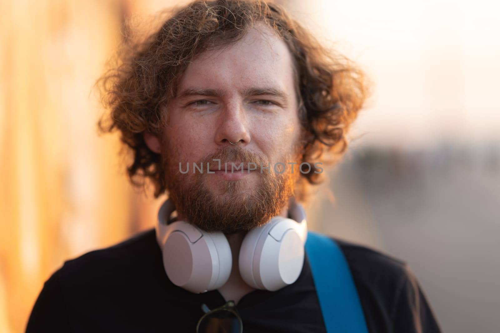 Bearded man hipster with wireless headphones on his neck standing outdoors at sunset. Portrait