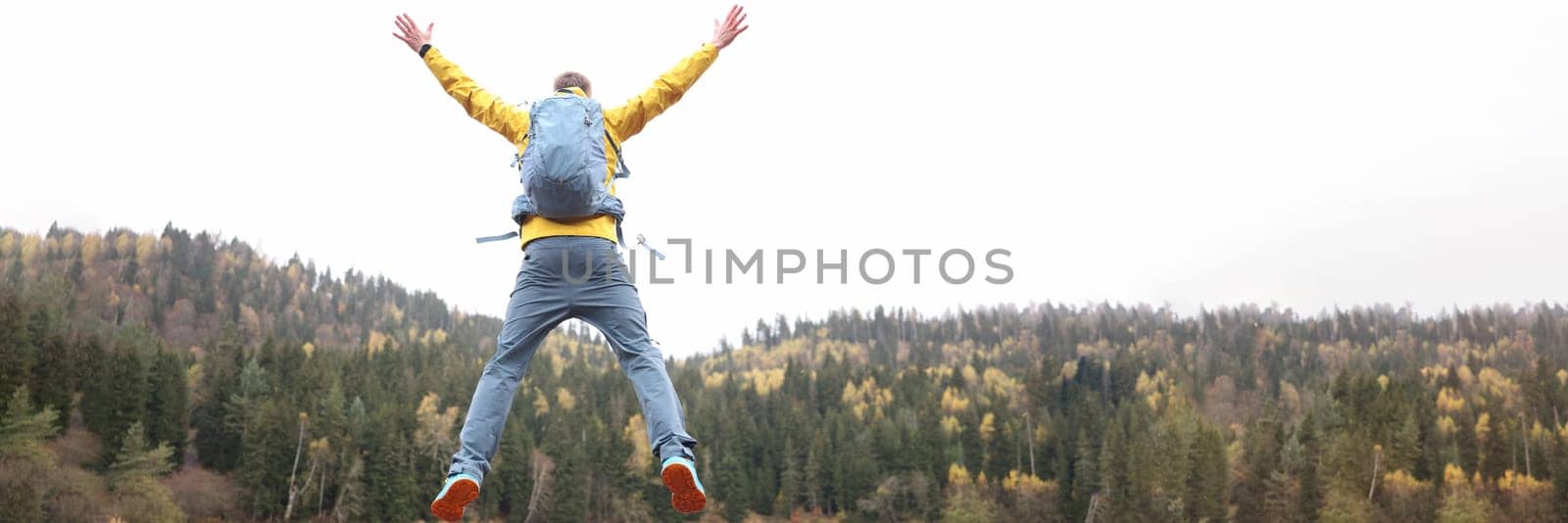 Happy male traveler with backpack jumping near forest lake back view. Travel and goal achievement concept