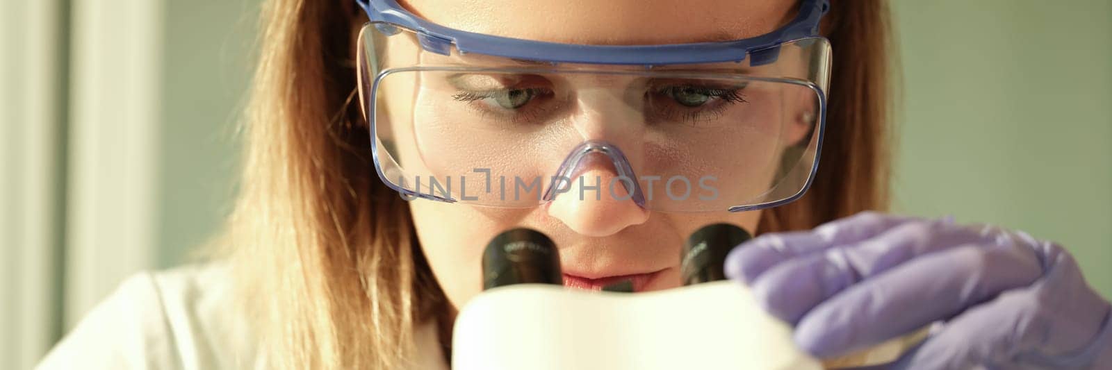 Woman scientist chemist in protective glasses looking through microscope in chemical lab. Laboratory diagnosis of internal diseases concept