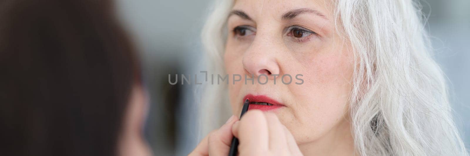 Cosmetologist painting lips with red lipstick to elderly woman by kuprevich