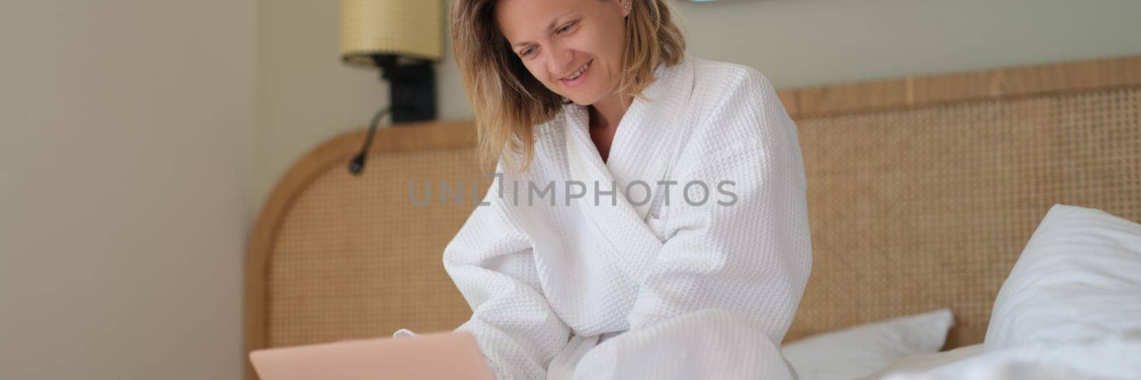 Young woman in white bathrobe working remotely with laptop in bed at home by kuprevich