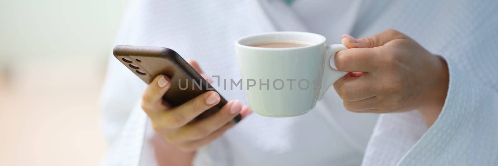 Woman in white bathrobe drinking coffee from cup and holding mobile phone at home closeup. Blogging social media concept