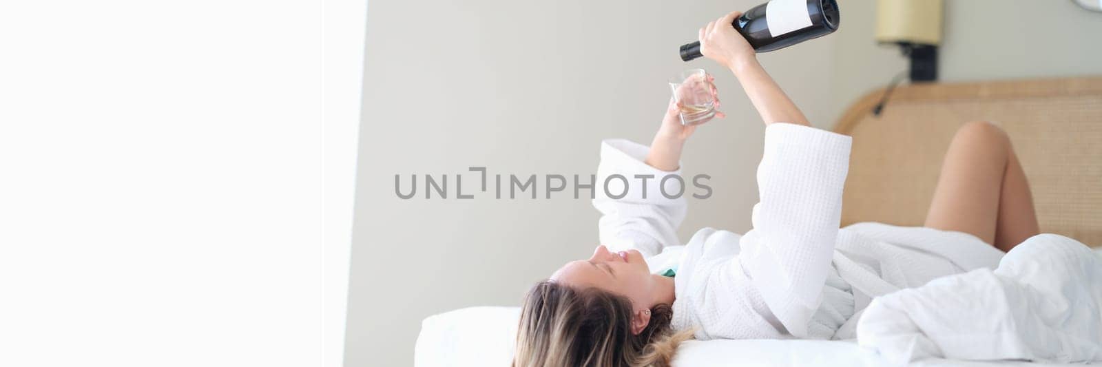 Woman in bathrobe lying in white bed and pouring champagne into glass. Rest and relaxation with alcohol in hotel concept