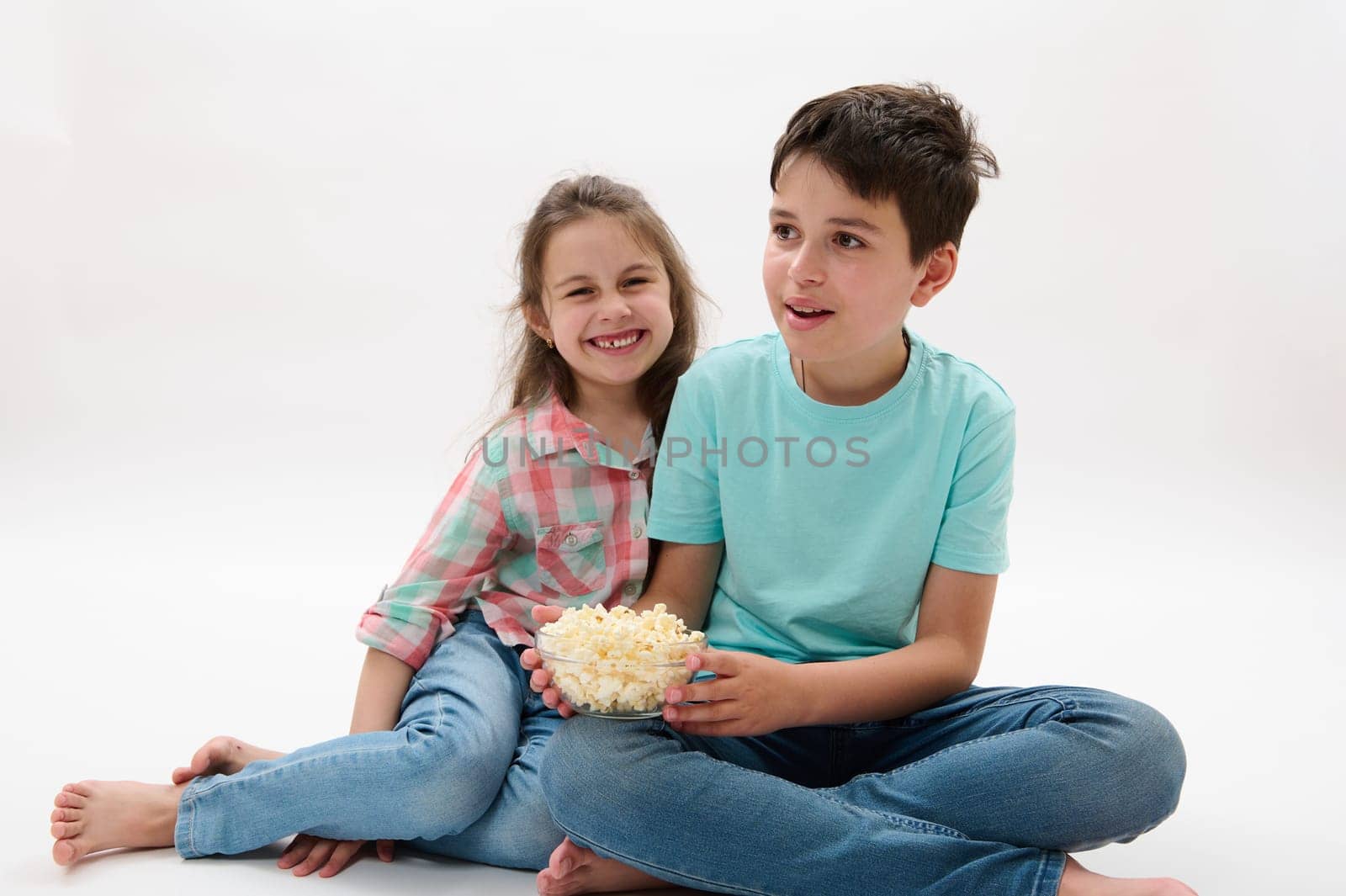 Mischievous adorable children, a teen boy and preschooler girl have fun together, eat popcorn and watch movie or cartoon by artgf