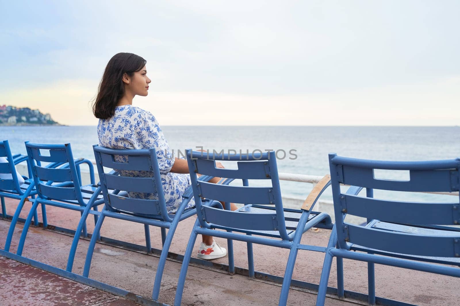 Beautiful smiling young mixed race woman sitting at one of the famous blue chairs at the Promenade des Anglais in Nice, France by berezko