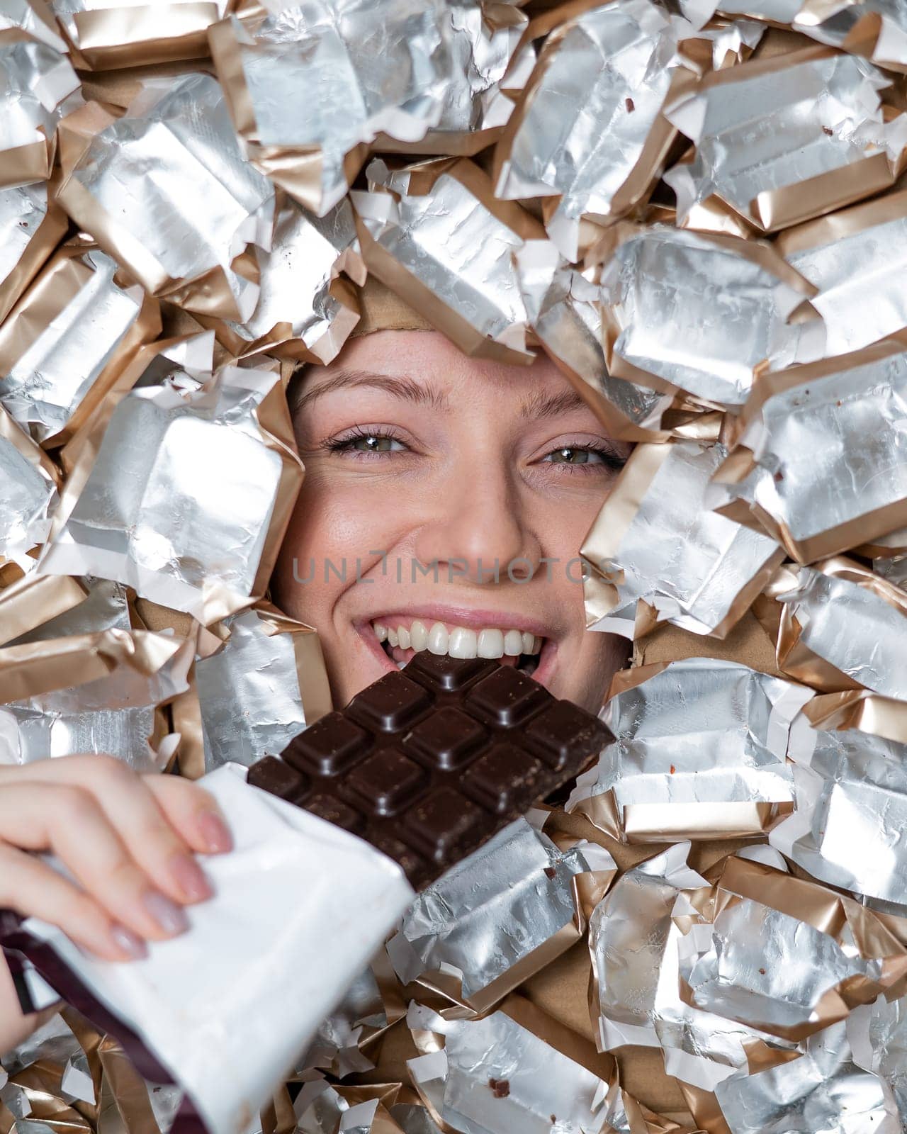 The face of a Caucasian woman surrounded by candy wrappers. The girl eats a bar of chocolate