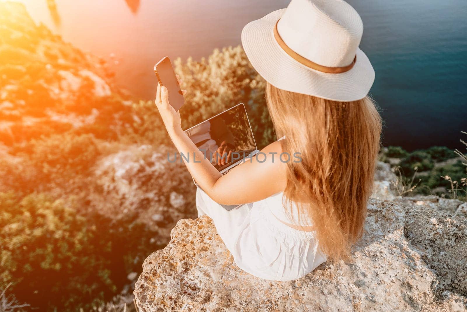 Successful business woman in yellow hat working on laptop by the sea. Pretty lady typing on computer at summer day outdoors. Freelance, travel and holidays concept.