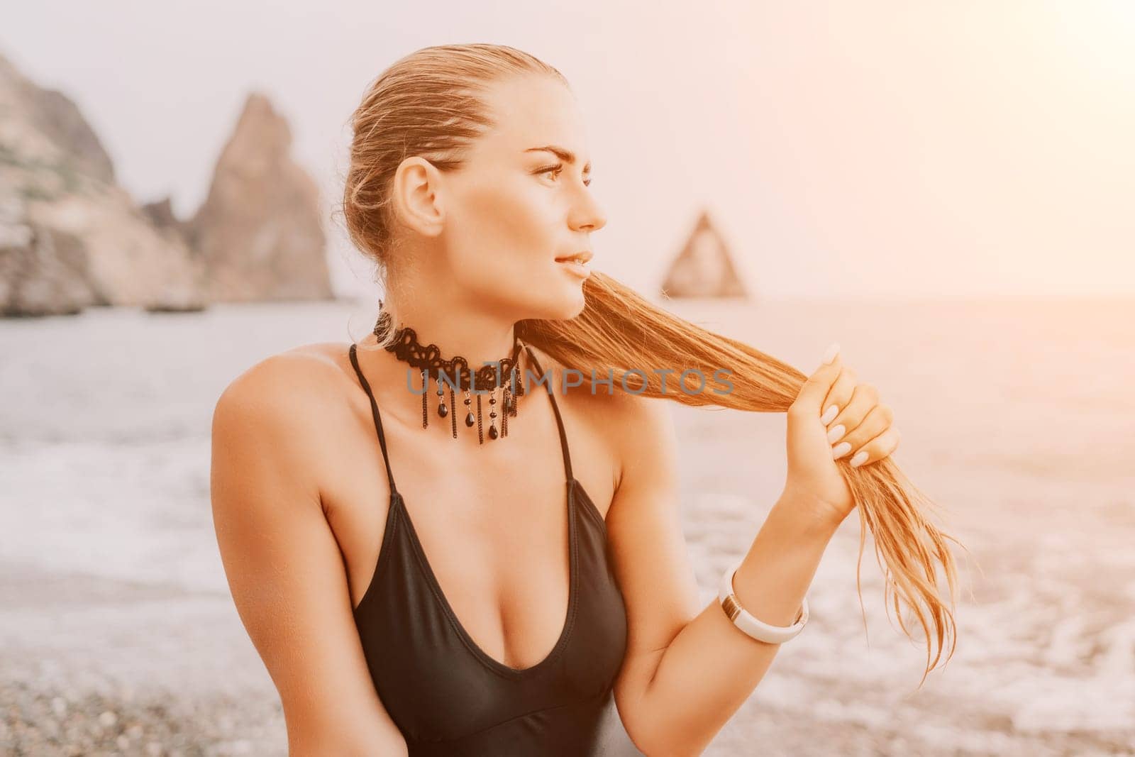 Woman travel portrait. Happy woman with long hair poses on a red volcanic rock at the beach. Close up portrait cute woman in black bikini, smiles at the camera, with the sea in the background