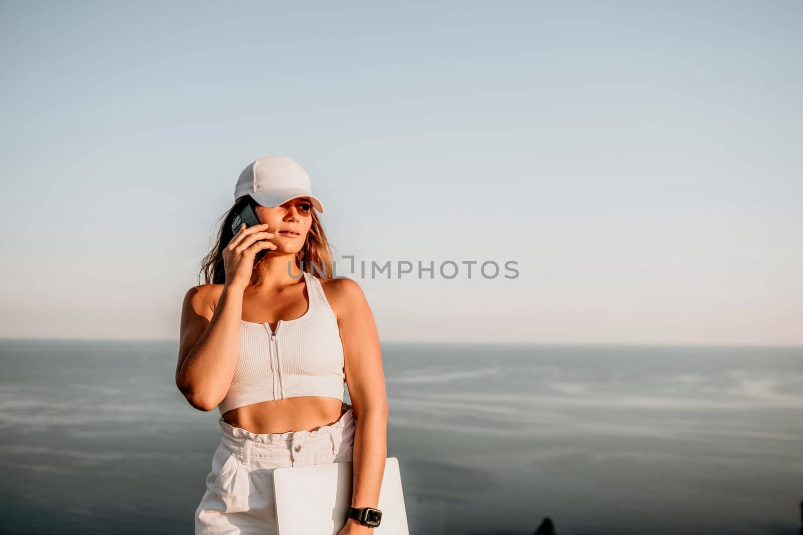 Successful business woman in yellow hat working on laptop by the sea. Pretty lady typing on computer at summer day outdoors. Freelance, travel and holidays concept.