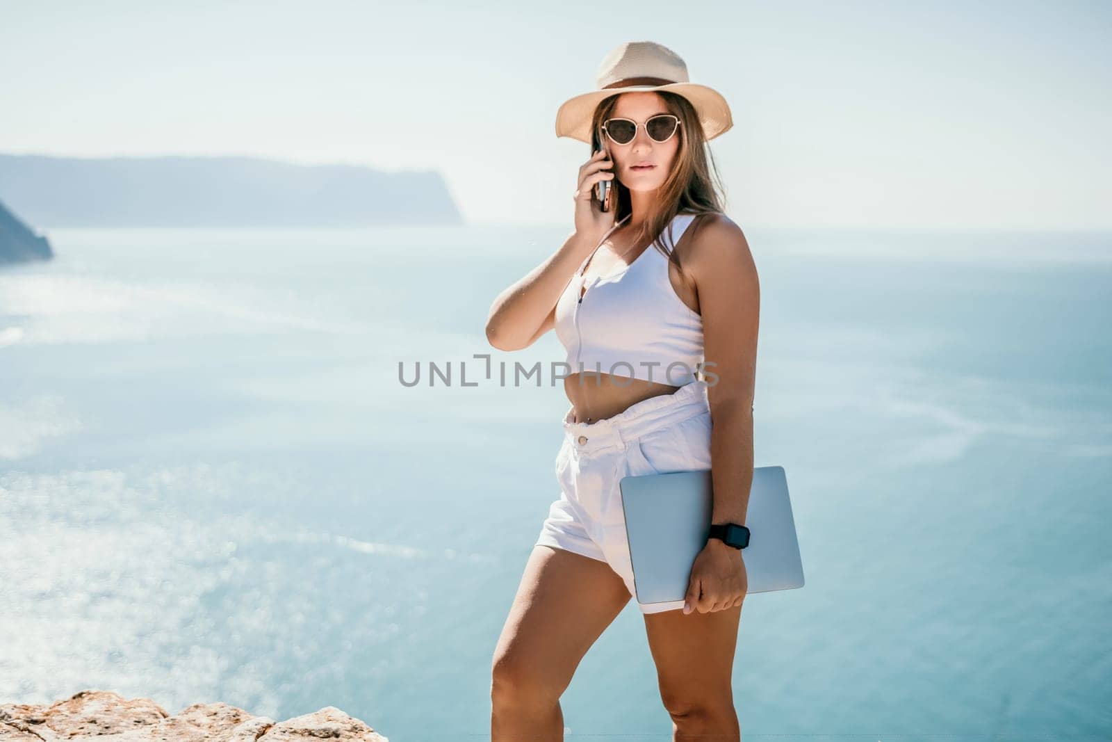 Successful business woman in yellow hat working on laptop by the sea. Pretty lady typing on computer at summer day outdoors. Freelance, travel and holidays concept.