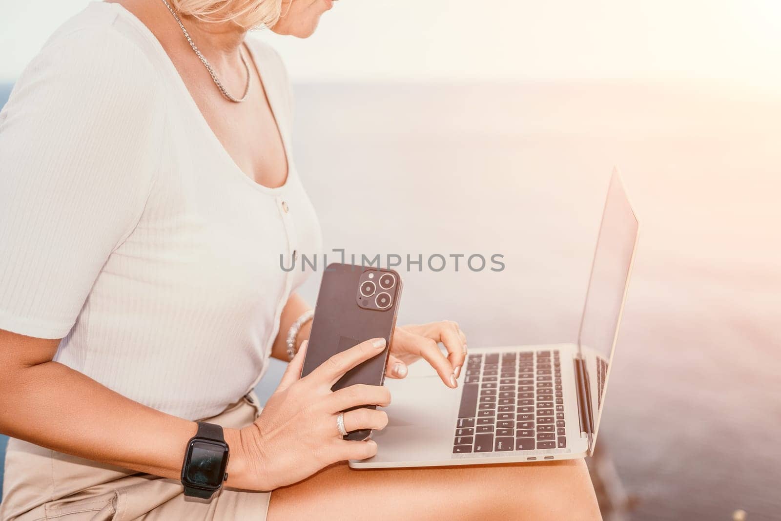 Digital nomad, Business woman working on laptop by the sea. Pretty lady typing on computer by the sea at sunset, makes a business transaction online from a distance. Freelance, remote work on vacation by panophotograph