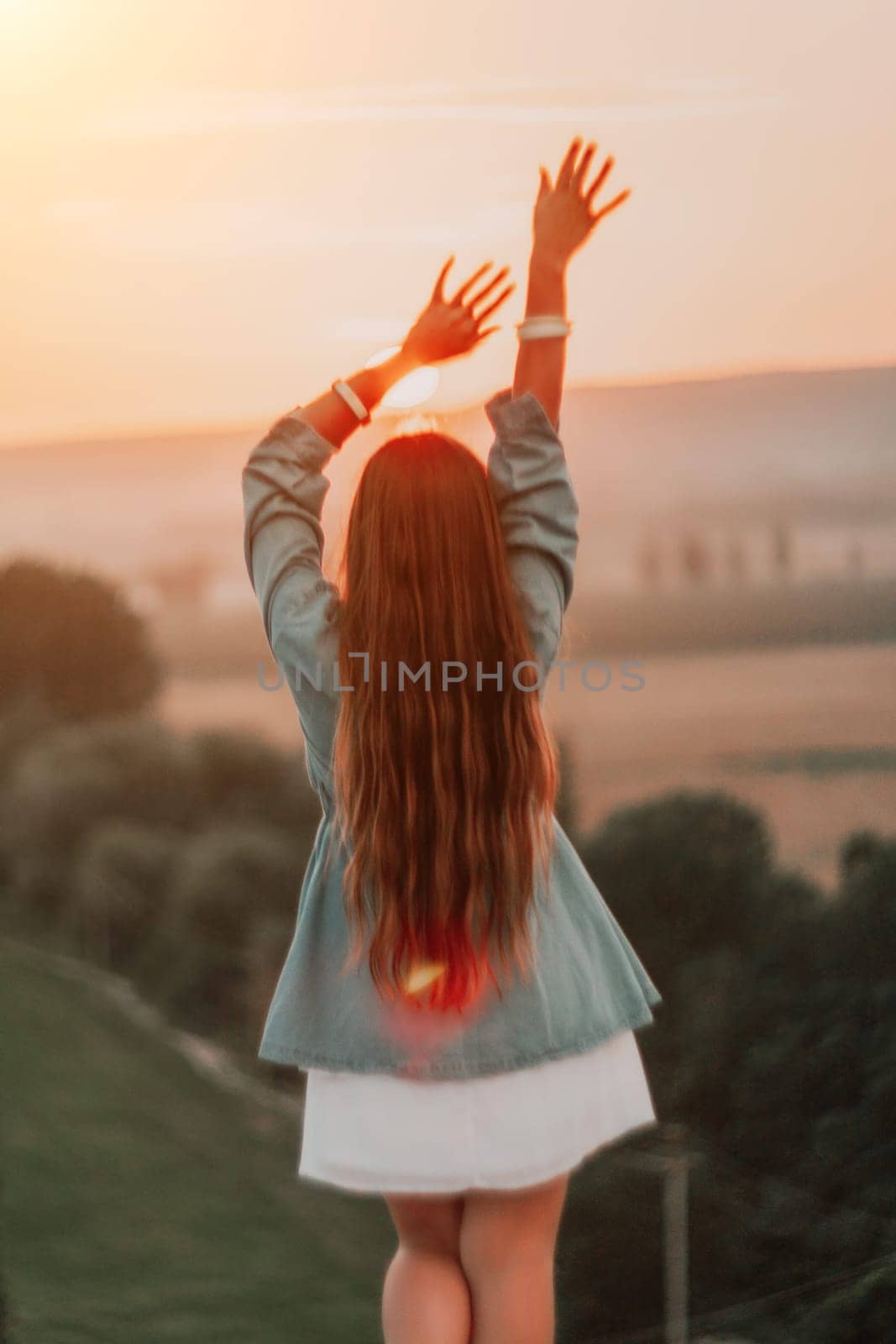 Happy woman standing with her back on the sunset in nature in summer with open hands. Romantic beautiful bride in white boho dress posing with mountains on sunset by panophotograph