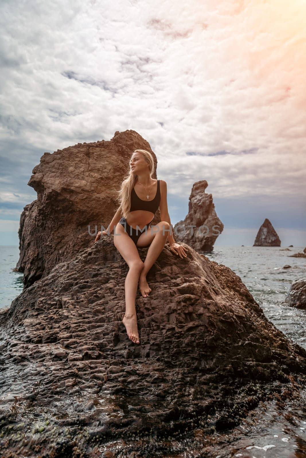 Woman travel sea. Attractive blonde woman in a black swimsuit enjoying the sea air on the seashore around the rocks. Travel and vacation concept. by Matiunina